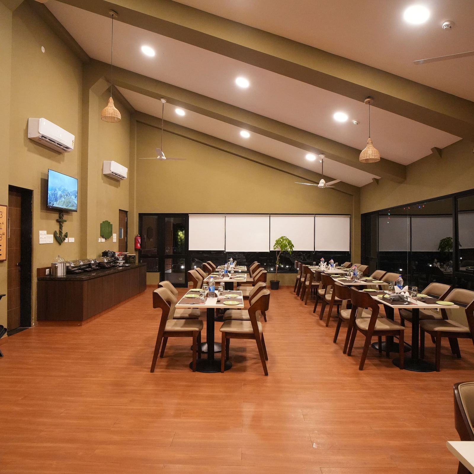 A restaurant with set tables and chairs, featuring a high, sloped ceiling and warm lighting. It includes a buffet area, air-conditioning units, and a wooden floor - Gaurav Lords Resort Shrivardhan.