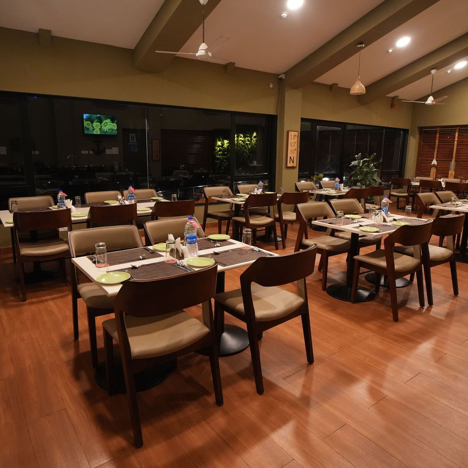 A quiet restaurant with dark wood tables and chairs, offering a warm, dimly lit ambience. It features large windows, wooden floors, and potted plants - Gaurav Lords Resort Shrivardhan.