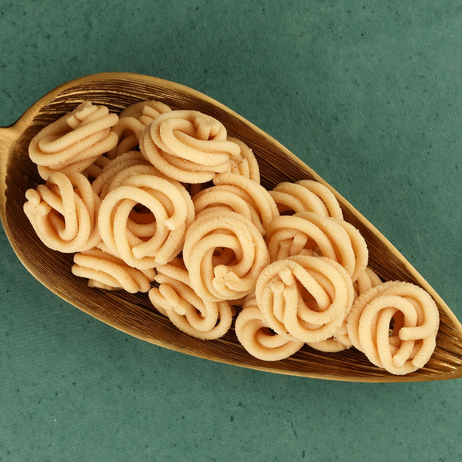 Golden, crispy, spiral-shaped Indian snack in a decorative scoop against a teal background.