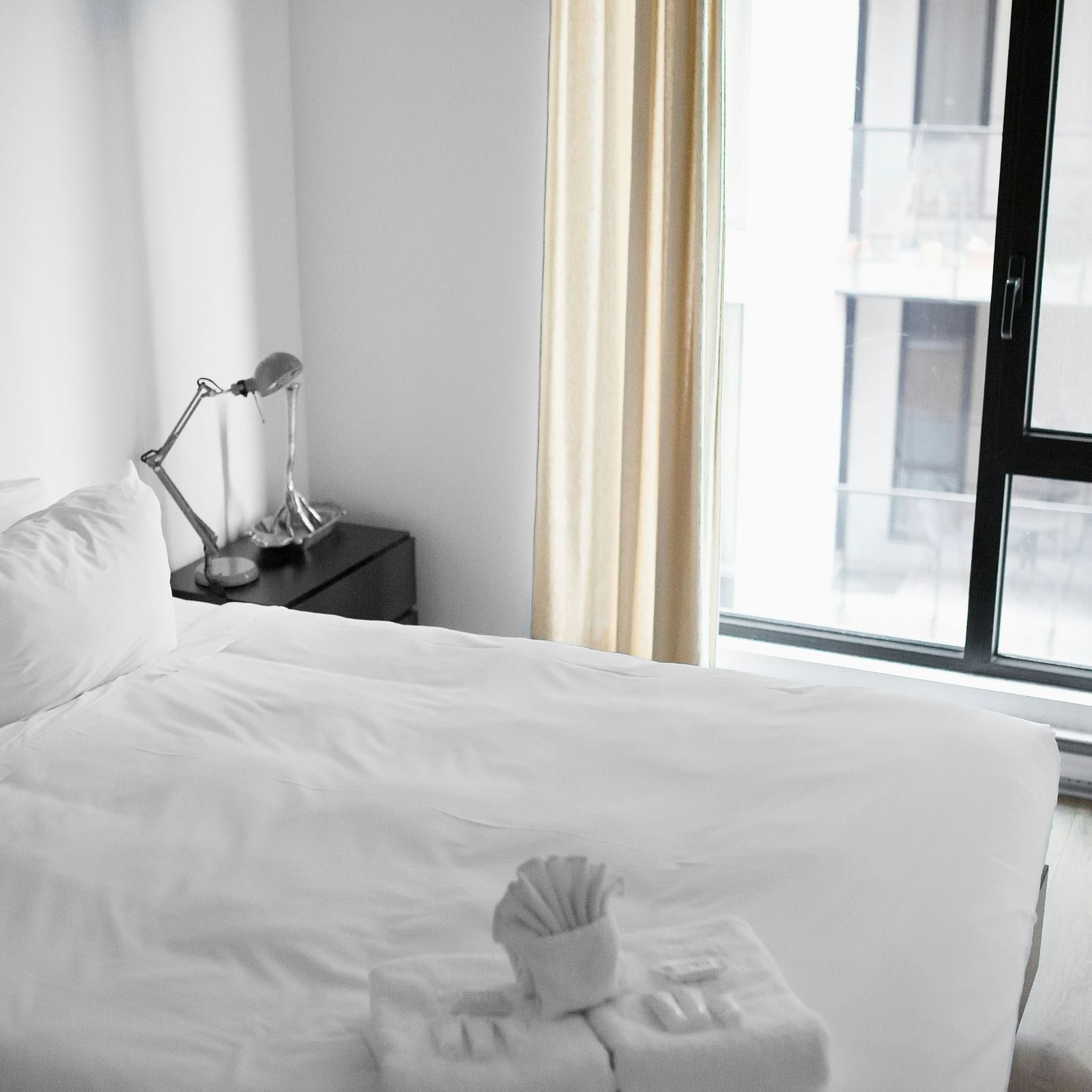 A hotel room with white sheets, side table and lamp next to the window