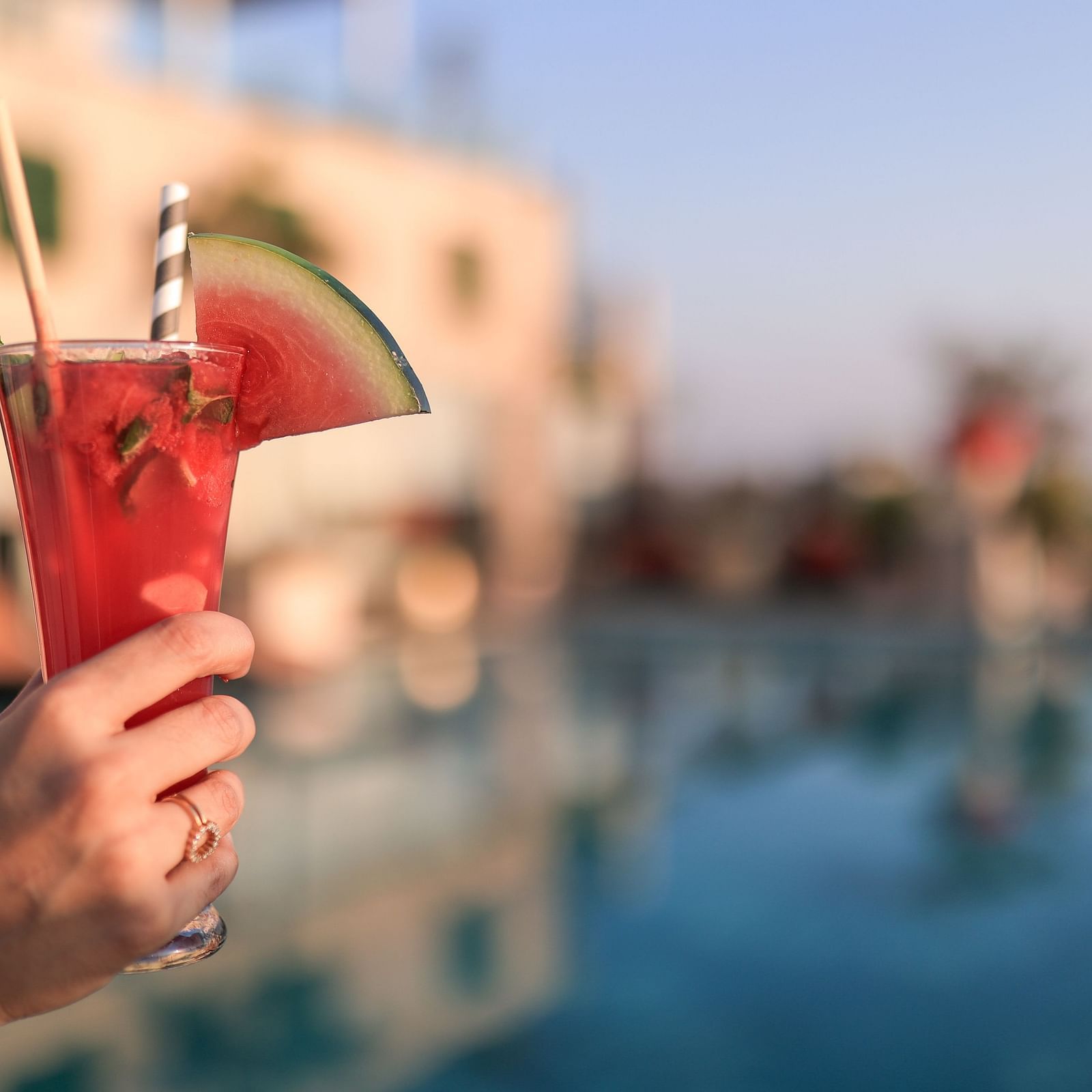 A person's hand holding a tall glass of watermelon juice, garnished with a slice of watermelon and a straw, against a blurred background.