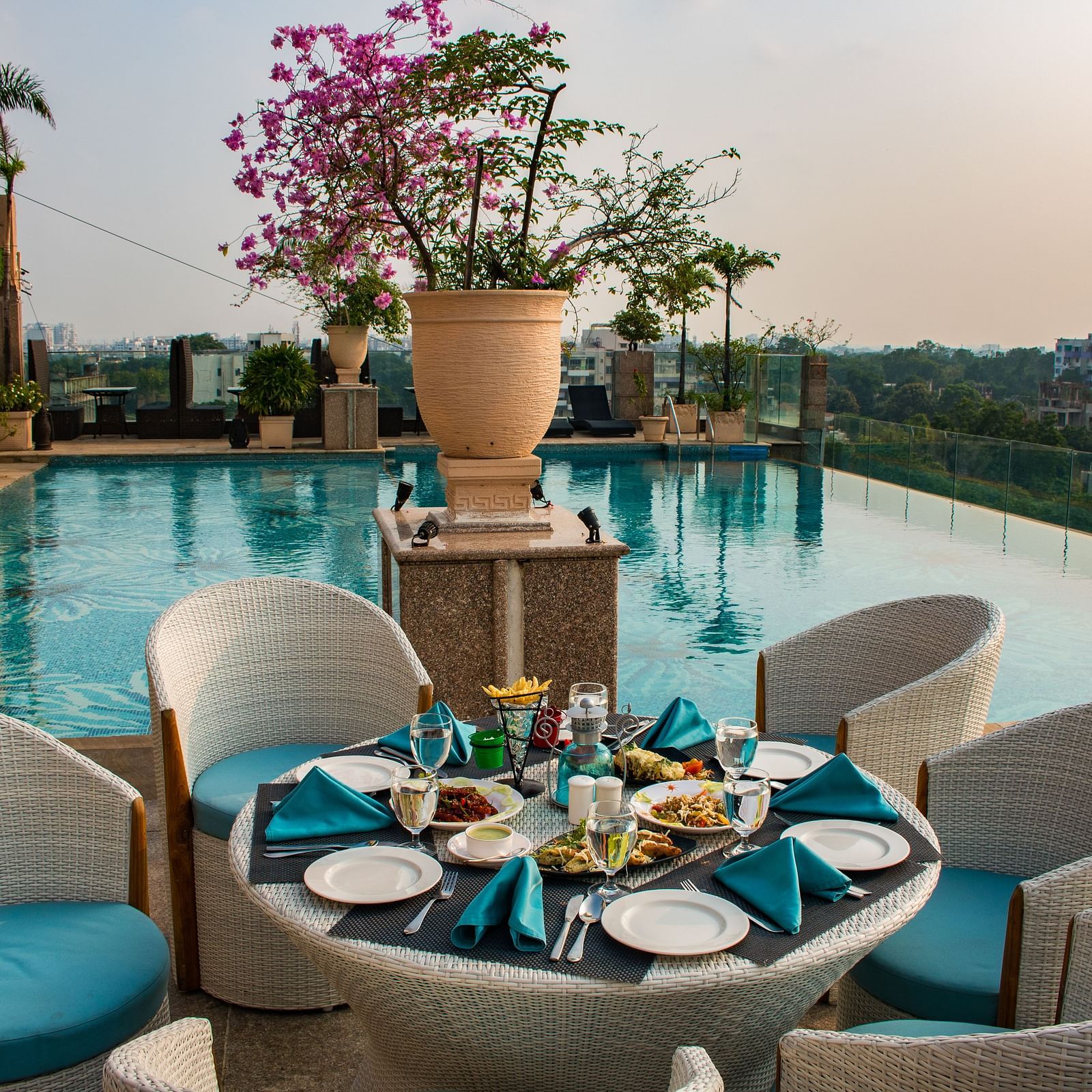 An outdoor dining at Hotel Ashok Nagpur featuring a round table with place settings for four, surrounded by cushioned chairs, and a swimming pool and cityscape in the background. 