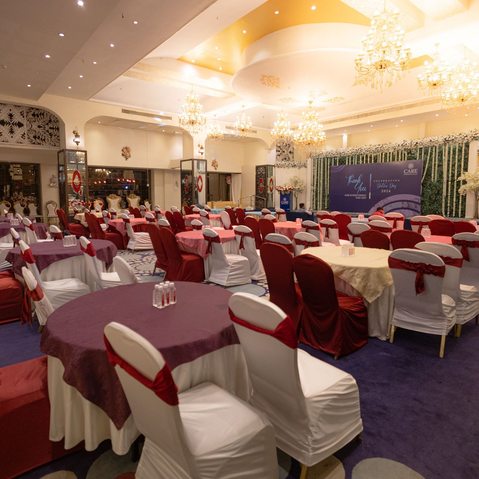 A banquet hall setup at Hotel Ashok Nagppur with tables draped in white linens and red accents, surrounded by chairs, and a stage area in the background.