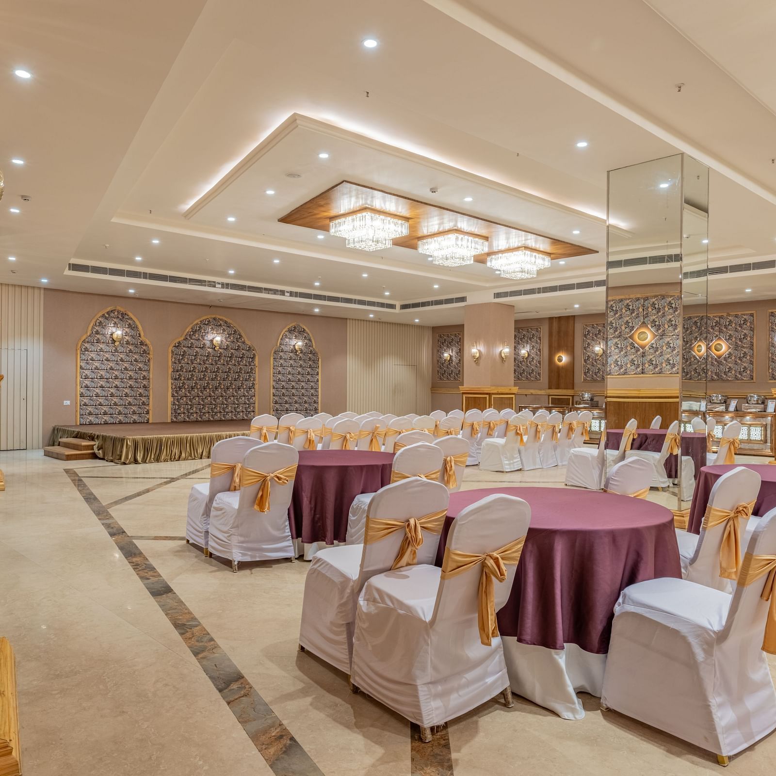 Banquet hall at Hotel Highway King, Bagru, with round tables, decorated chairs, chandeliers, and decorative wall panels.