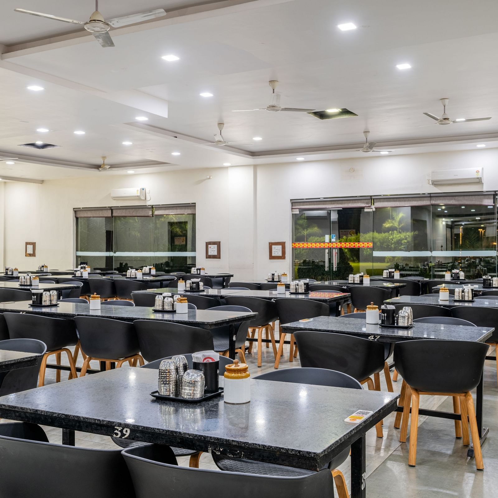 Extended view of the restaurant at Hotel Highway King, Behror, furnished with dining tables with varying seating capacities, ample lighting and windows that offer views of the trees outside.