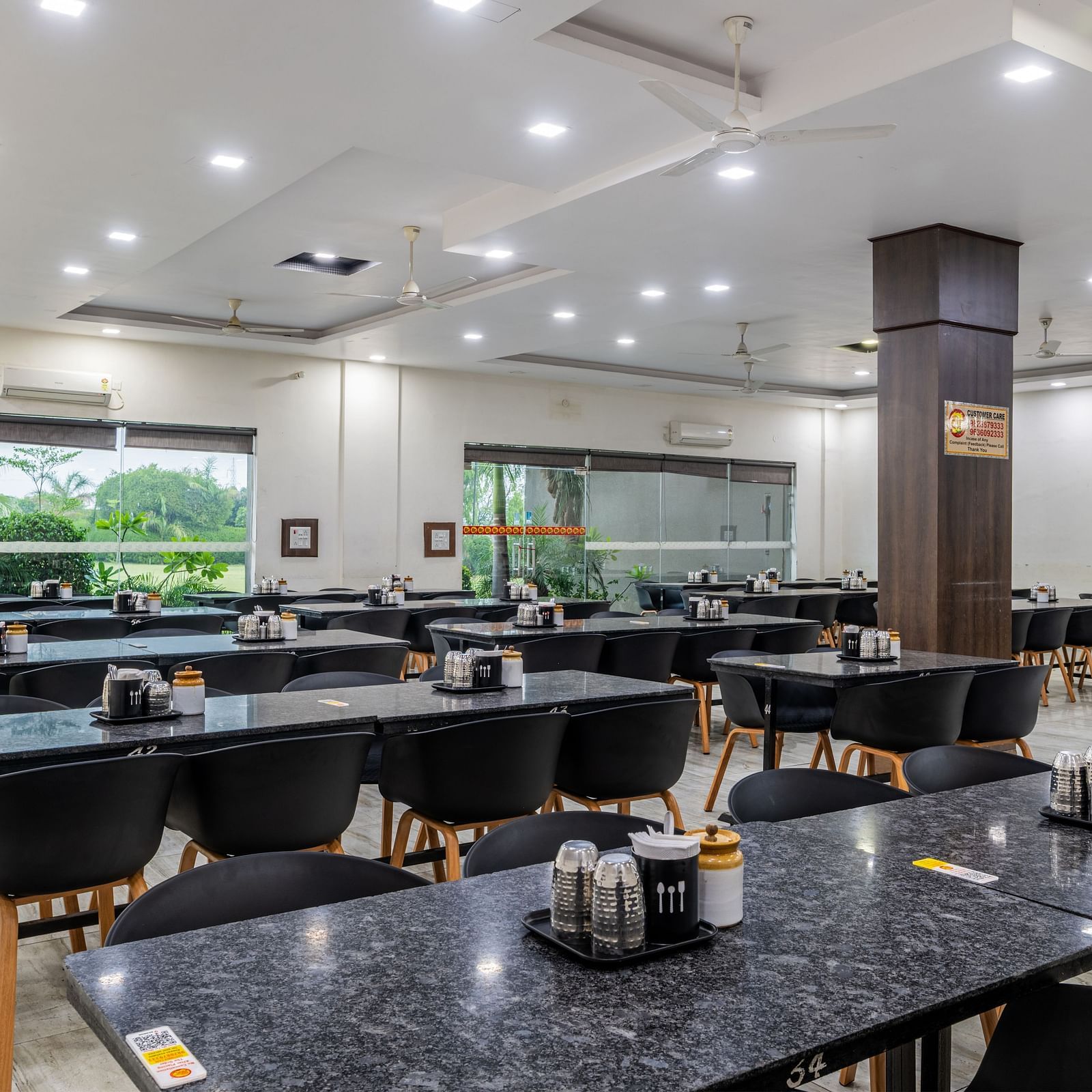 Extended view of the restaurant at Hotel Highway King, Behror, furnished with dining tables with varying seating capacities, ample lighting and windows that offer views of the trees outside.