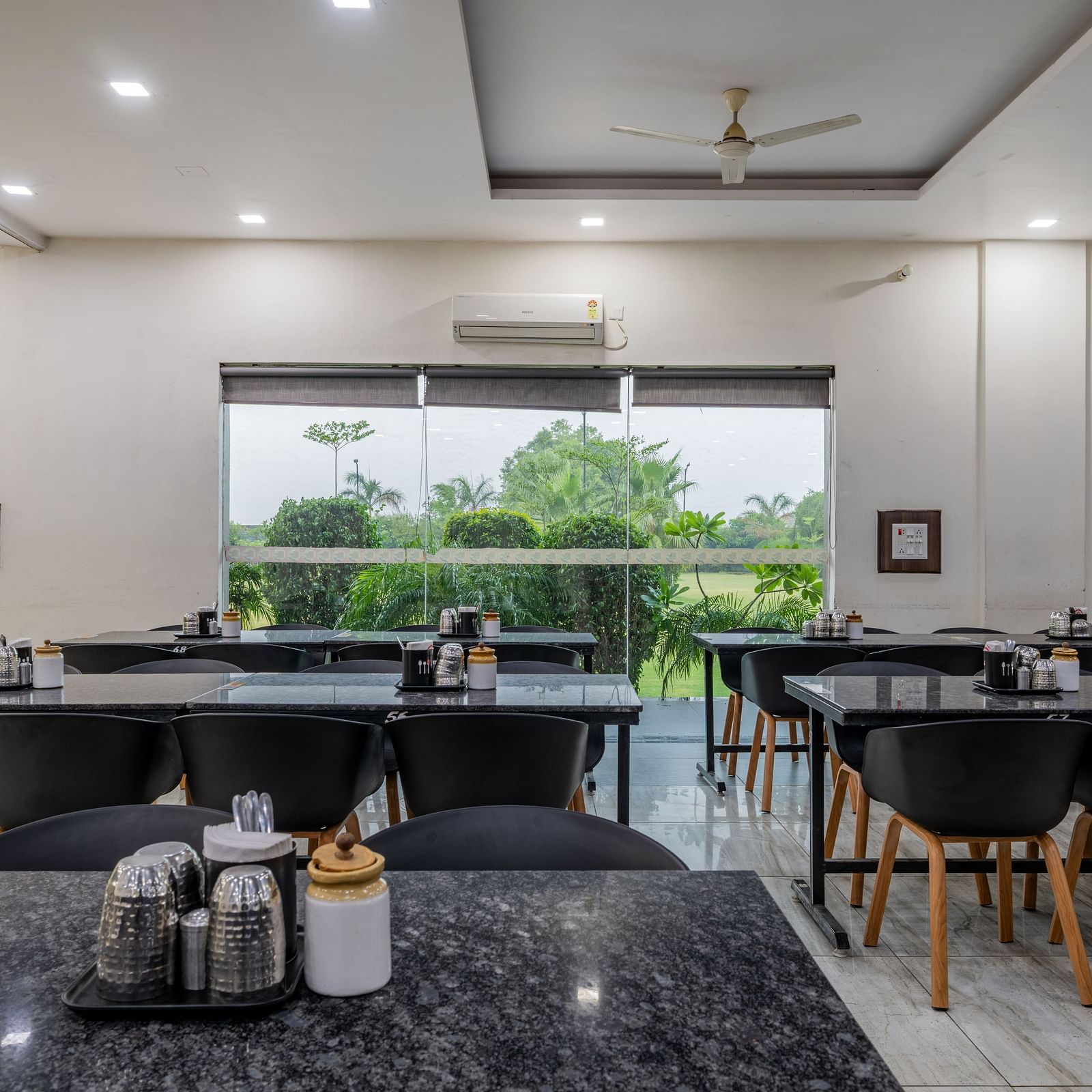 Front view of the dining tables reflecting the overhead lights, and glass windows offering view of the trees outdoor at Hotel Highway King, Behror.