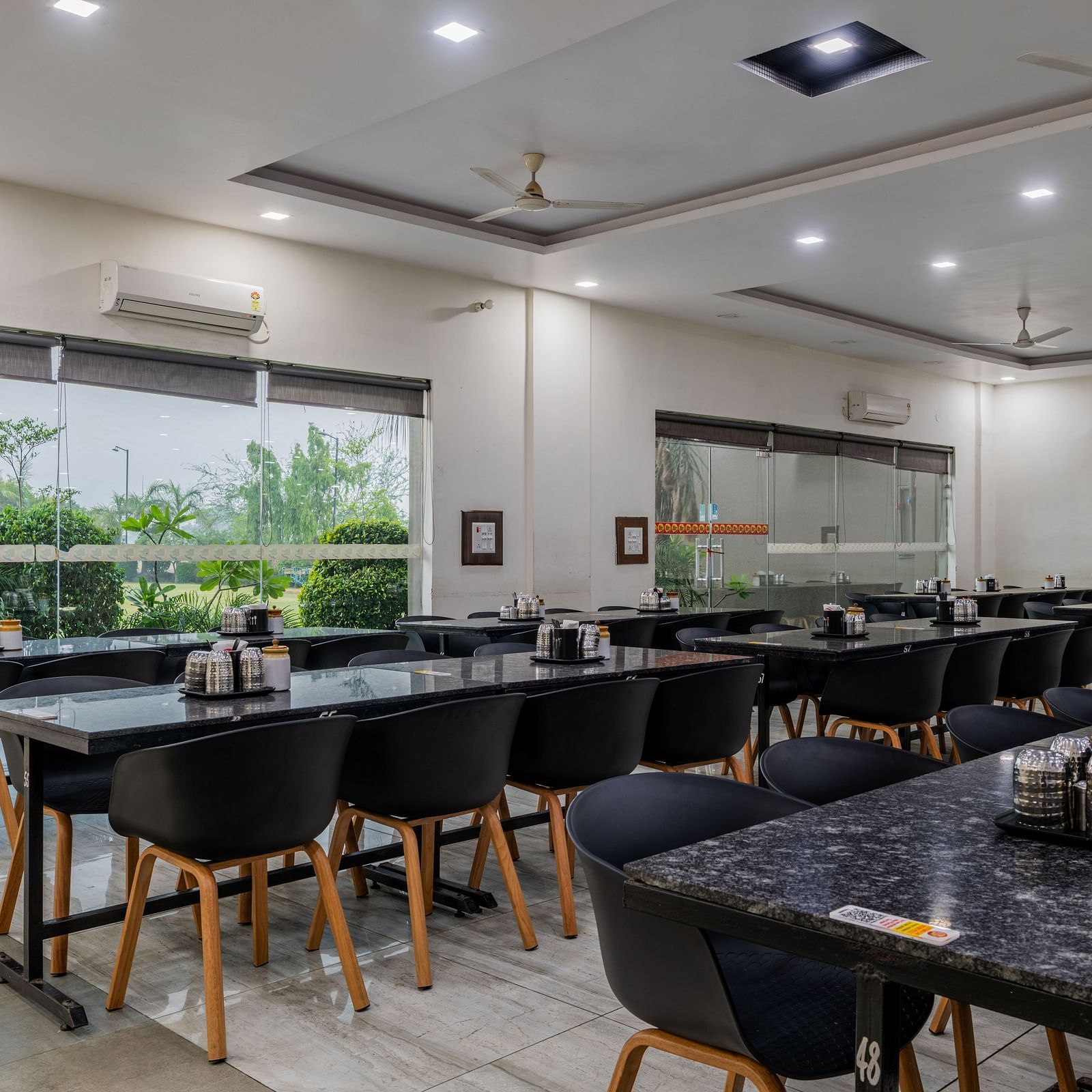 Air-conditioned restaurant of Hotel Highway King, Behror, is furnished with parallel aligned dining tables, water cups and glass windows that offer views of the trees outside at Hotel Highway King, Behror.