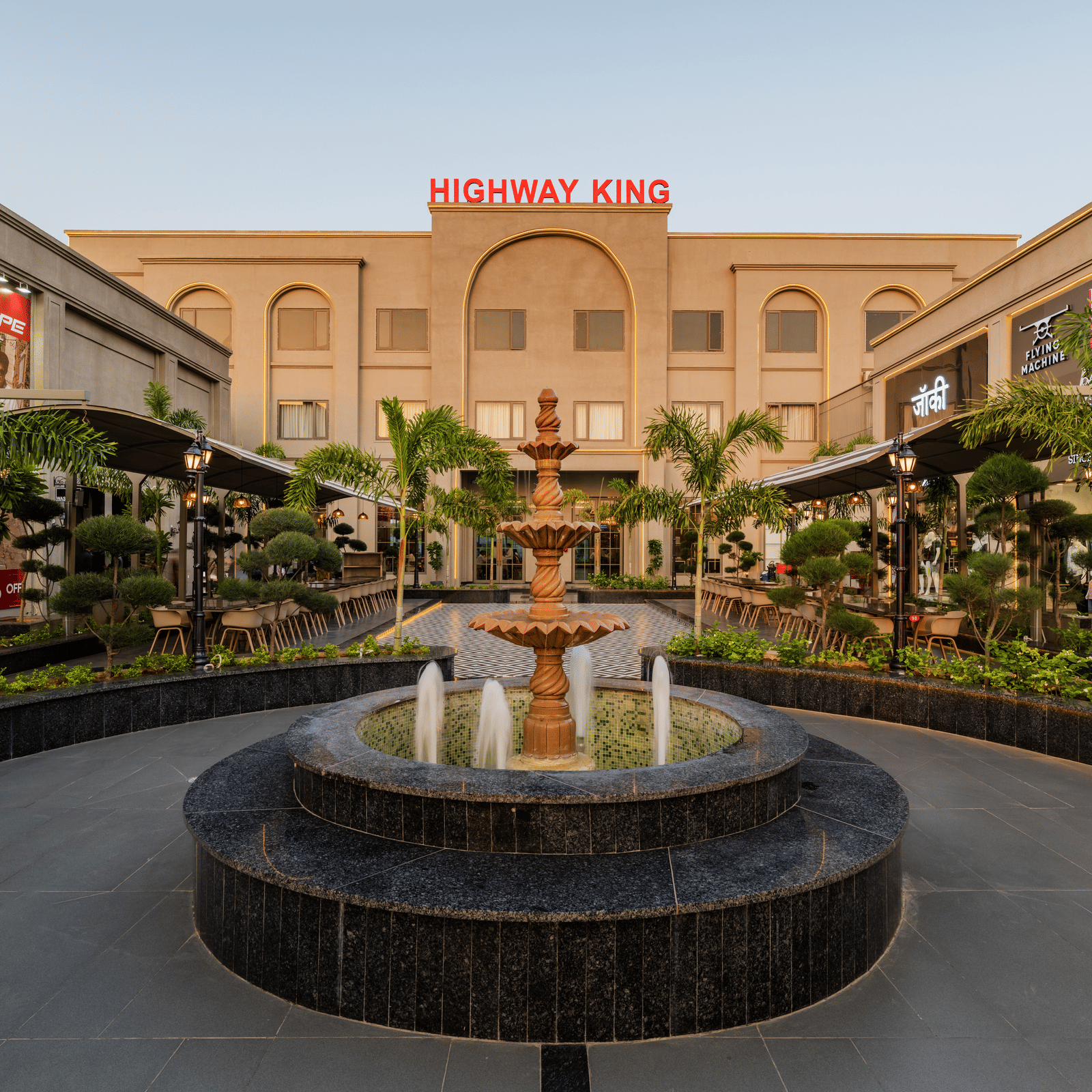 A central water fountain, surrounding storefronts, and a main building at Hotel Highway King, Harmada.