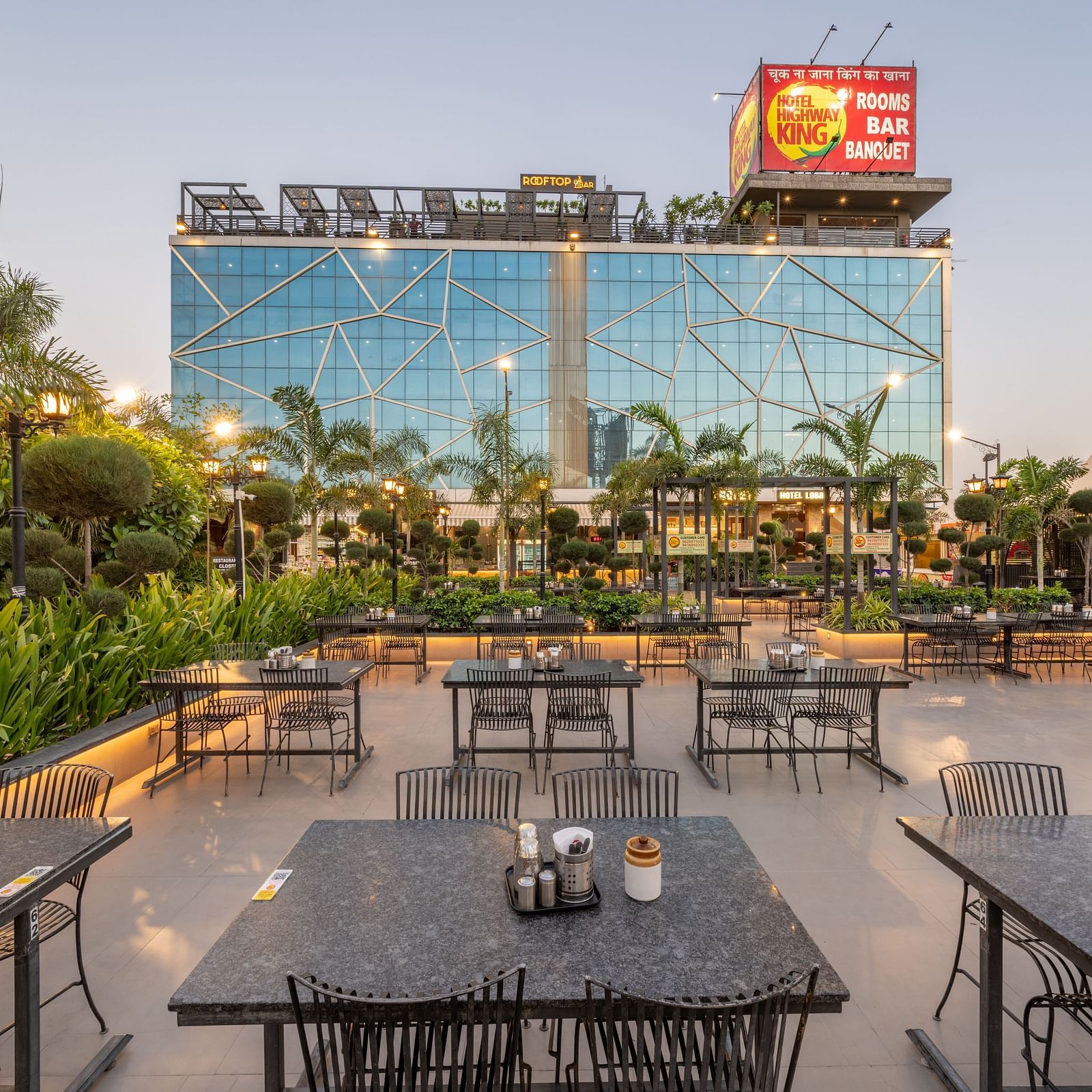 Outdoor seating area with multiple tables and chairs arranged in rows with greenery and the facade of Hotel Highway King at the background.