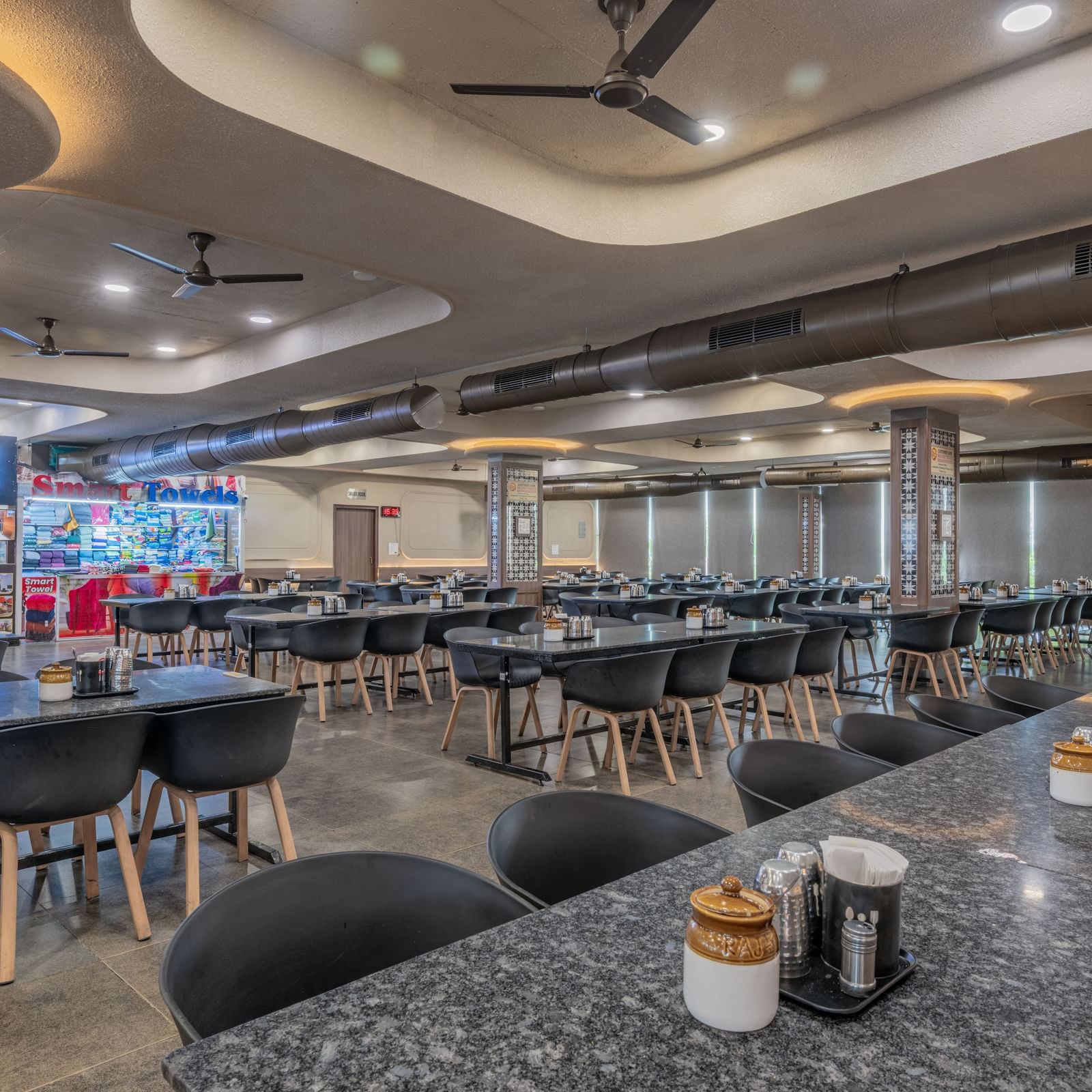 A restaurant interior with sleek tables, ambient ceiling lights, and neatly arranged seating at Hotel Highway King, Neelka.