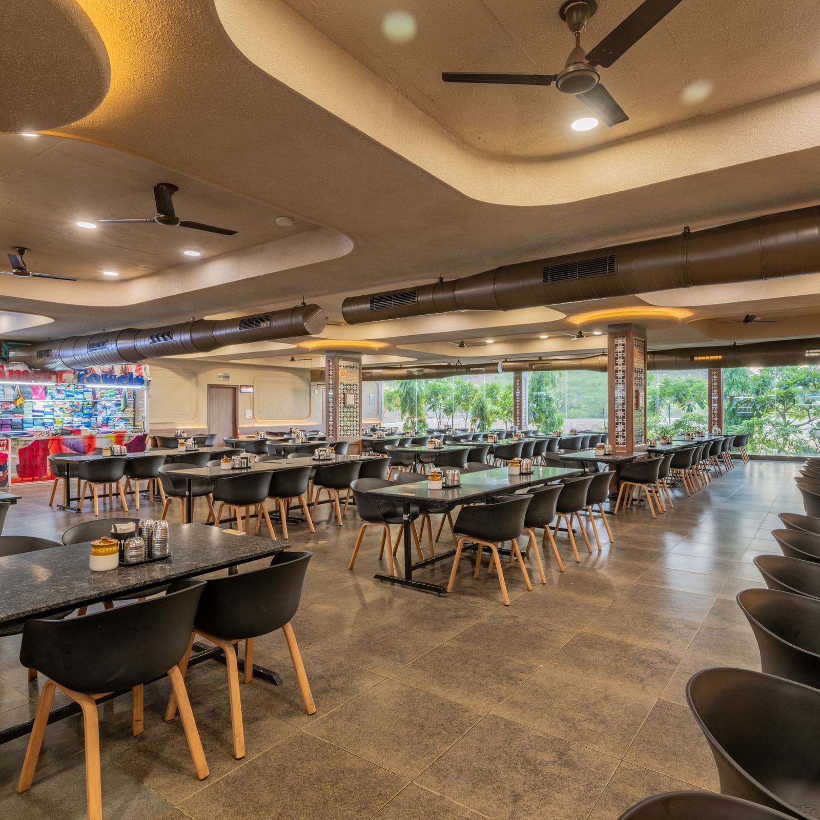 A diagonal view of the restaurant seating area, featuring panoramic glass panels in the background and warm lighting, at Hotel Highway King, Neelka.