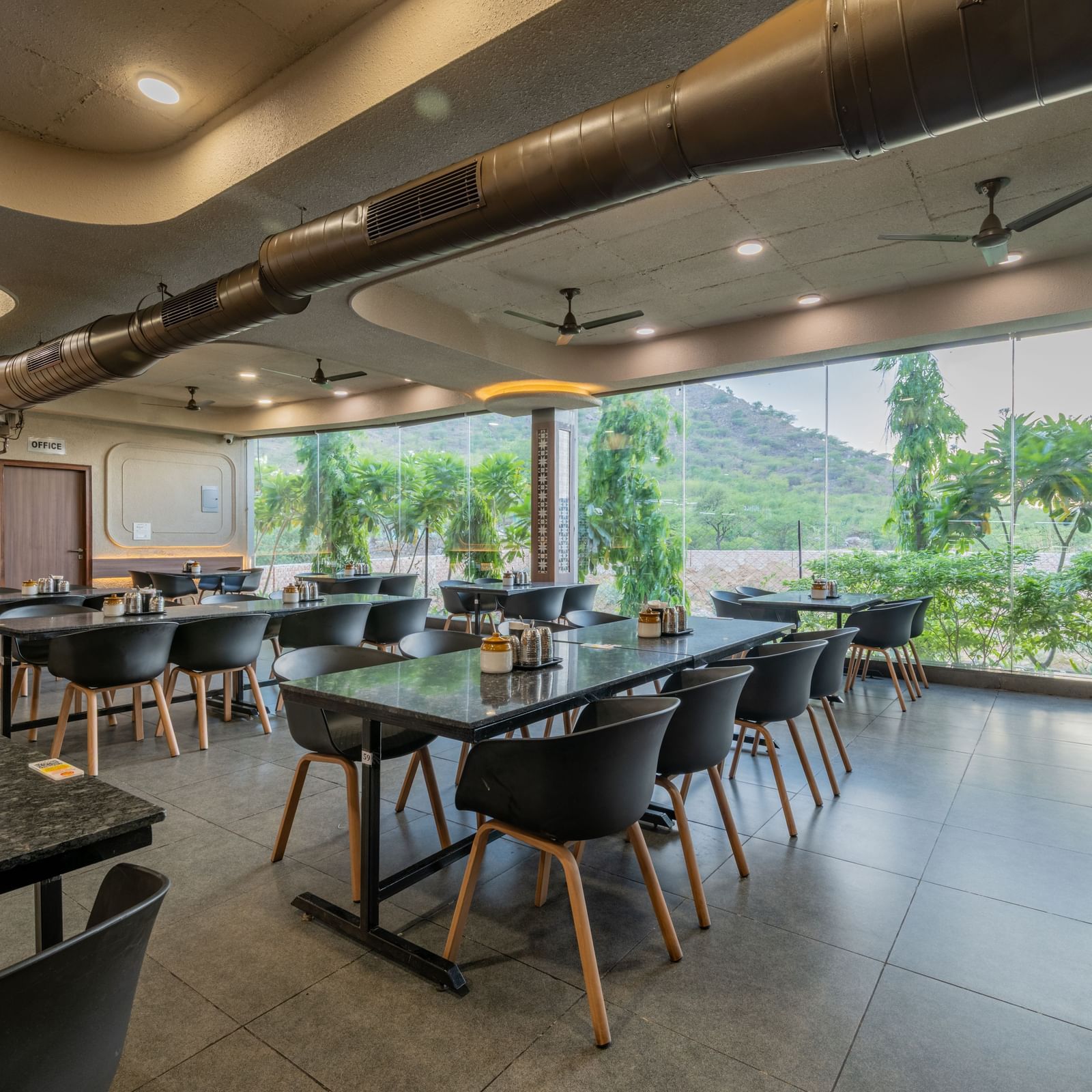 Diagonal view of the restaurant interior at Hotel Highway King, Neelka, with glass panels behind the seating area overlooking the lawn and lush mountains.