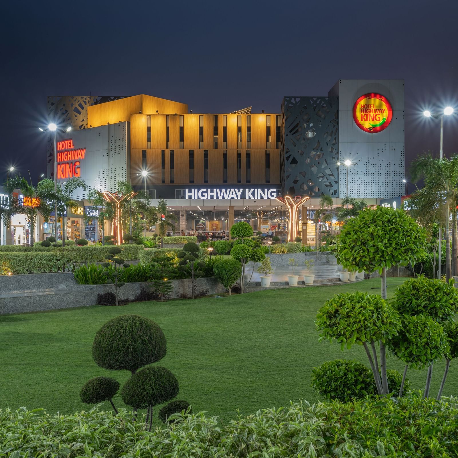 An illuminated modern building with a distinctive sign stands behind a well-maintained green lawn and manicured shrubbery at dusk.