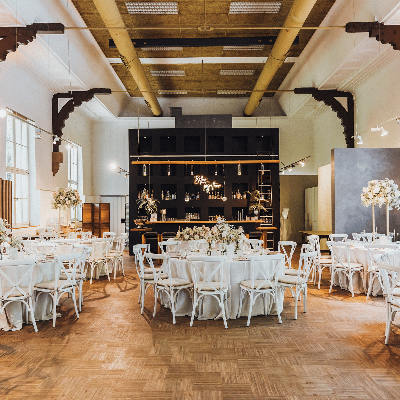 Elegantly set dining hall with white chairs, round tables with white linens and a central bar area under rustic industrial beams