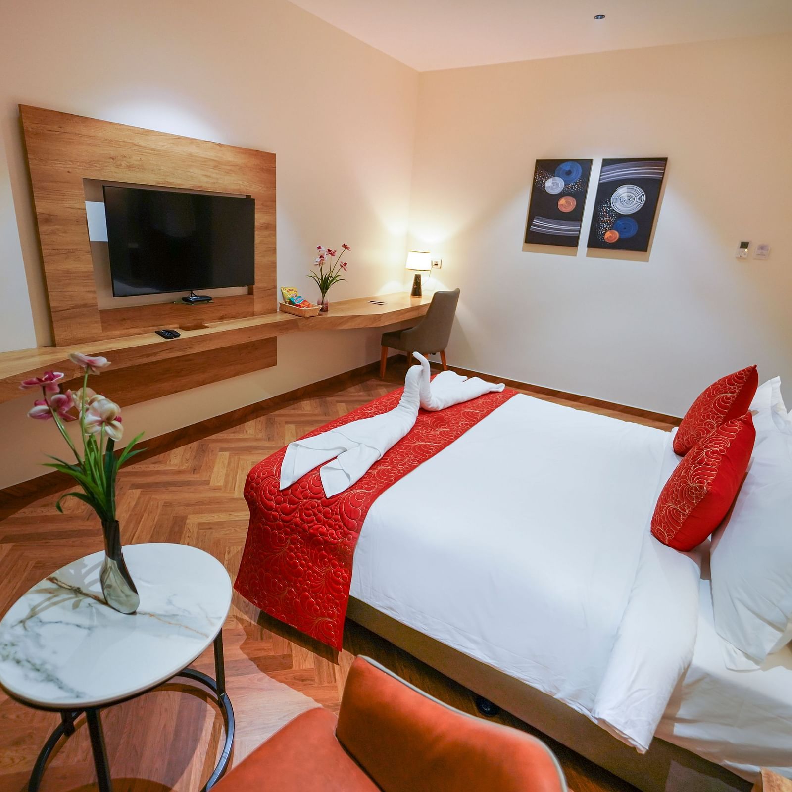 A hotel room features a bed with pillows and folded linens, a mounted television, a desk area, a small round table with flowers, and artwork on the wall - Hotel Park Inn, Nellore.