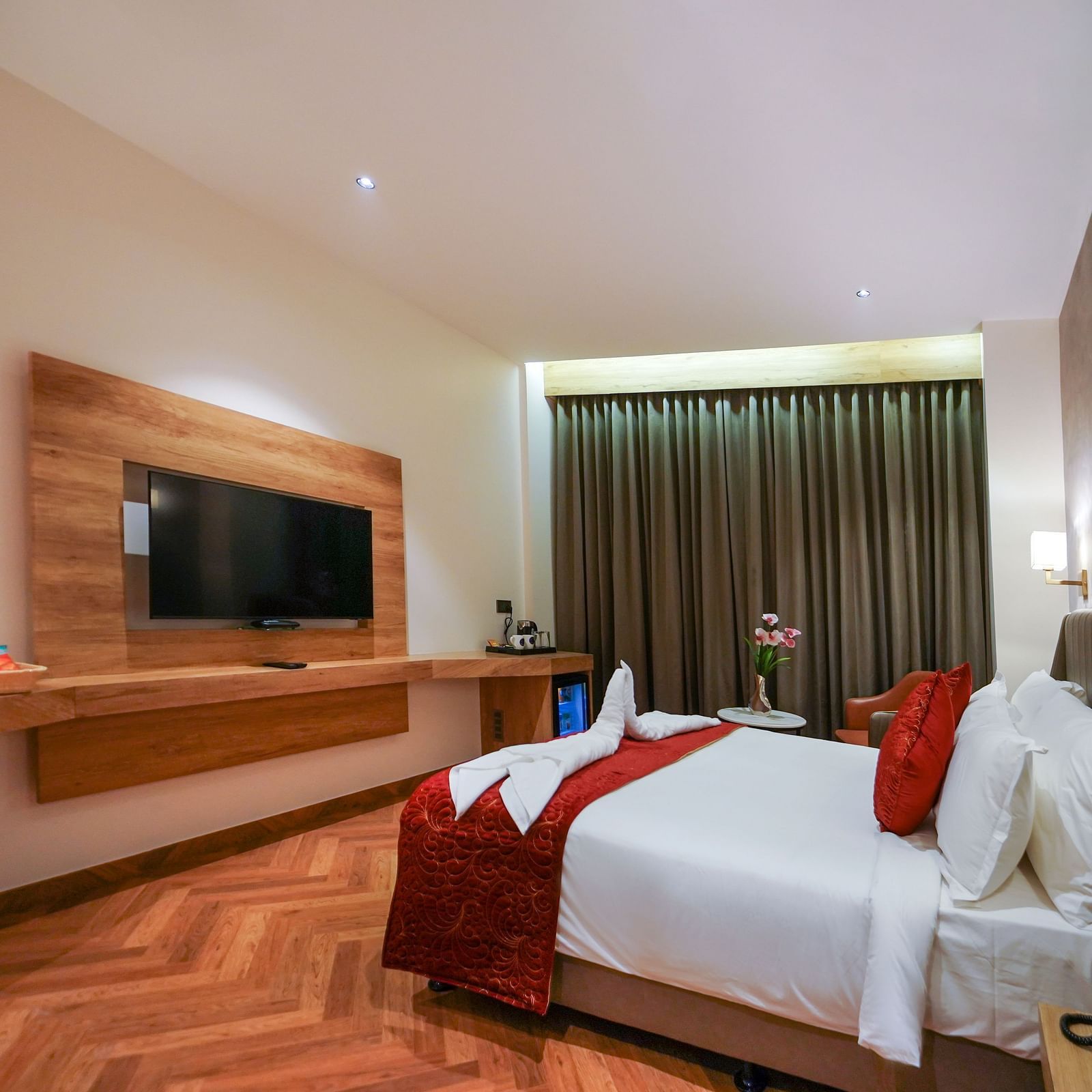 A hotel room contains a bed with pillows and decorative linens, a mounted television, a desk area with flowers, and window curtains - Hotel Park Inn, Nellore.
