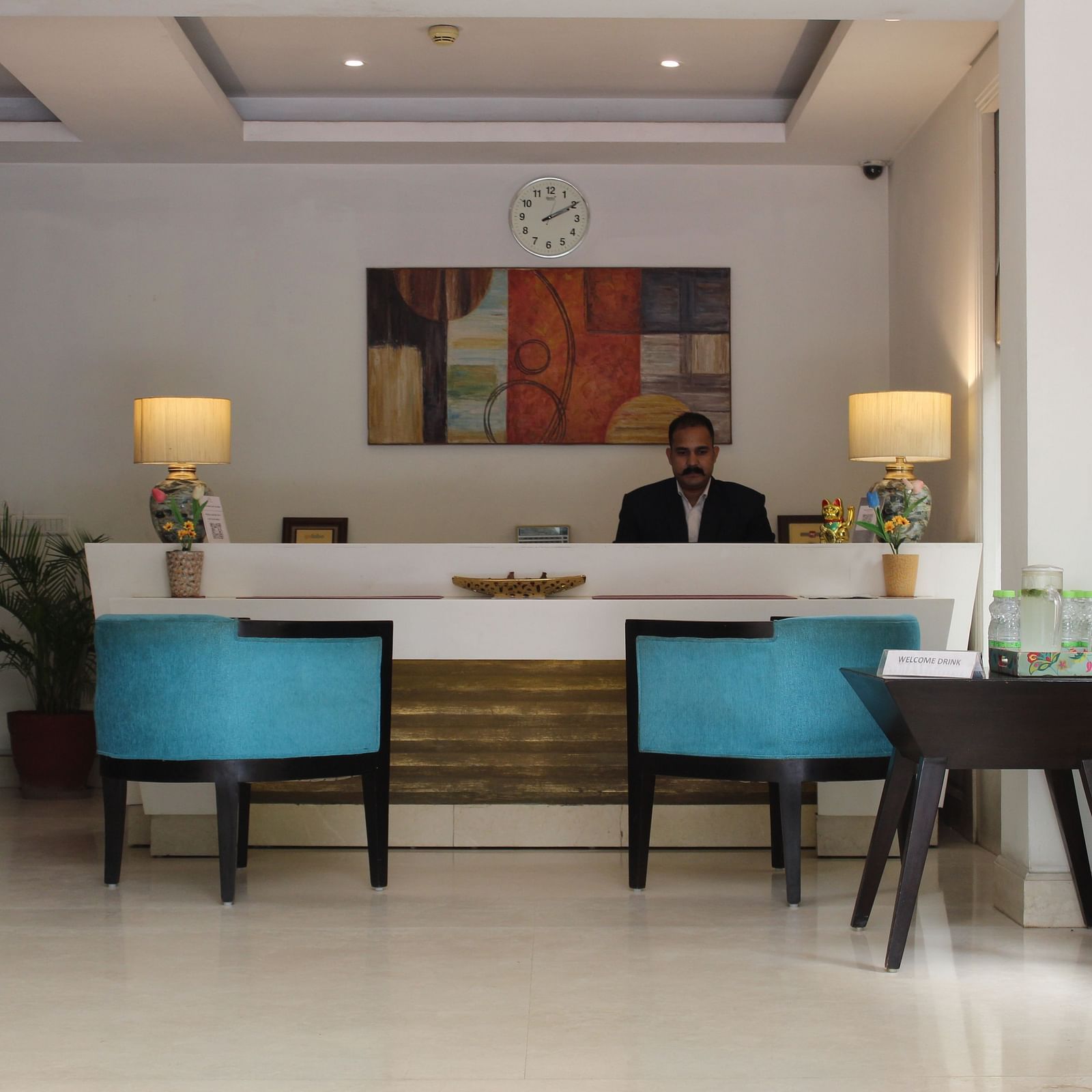 An image of a reception lobby featuring a reception desk where a reception is standing and in front of them there are two chairs placed along with a table at the corner of the room - Inde Hotels Vista Woods