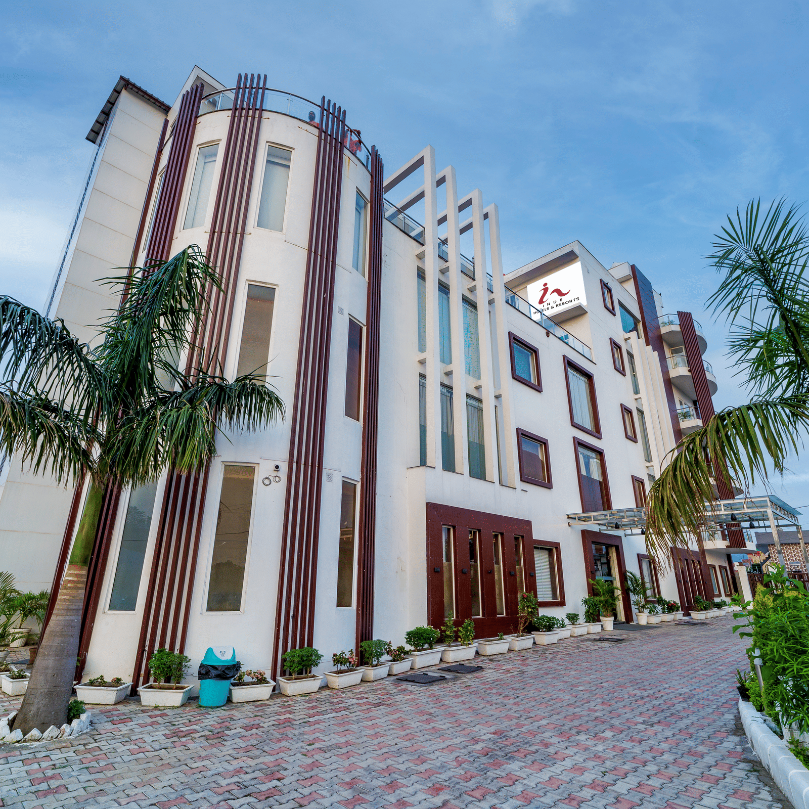 Exterior view of Inde Reva Resorts Rishhikesh showing the multi-storey building with palm trees, paved driveway, and lush green on the right.