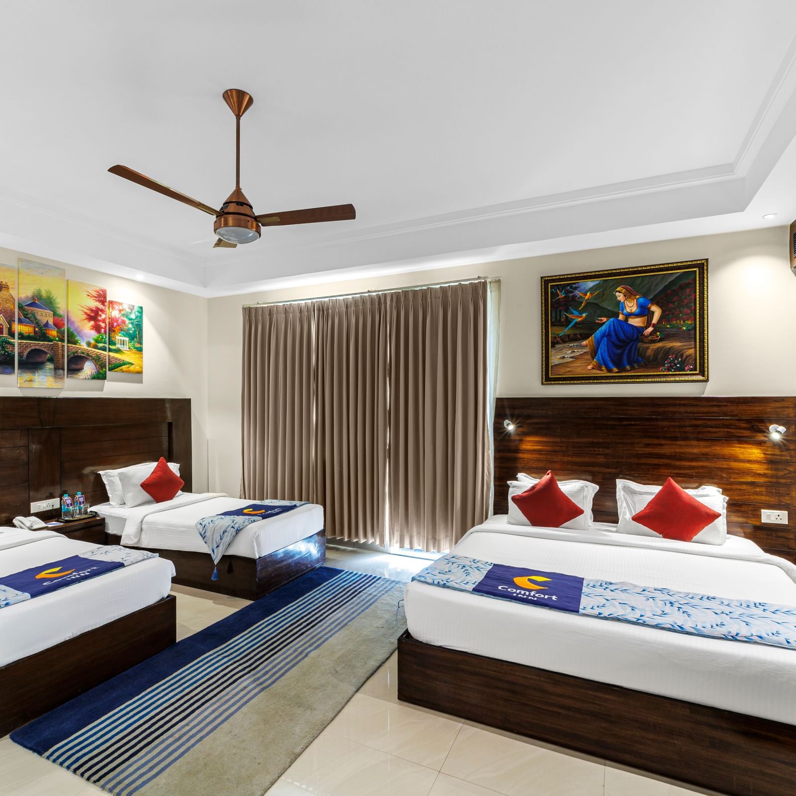 Well furnished huge room with a king size bed and twin beds featuring a ceiling fan, artwork above bedside table, and TV unit opposite the beds at Inder Reva Resorts Rishhikesh.