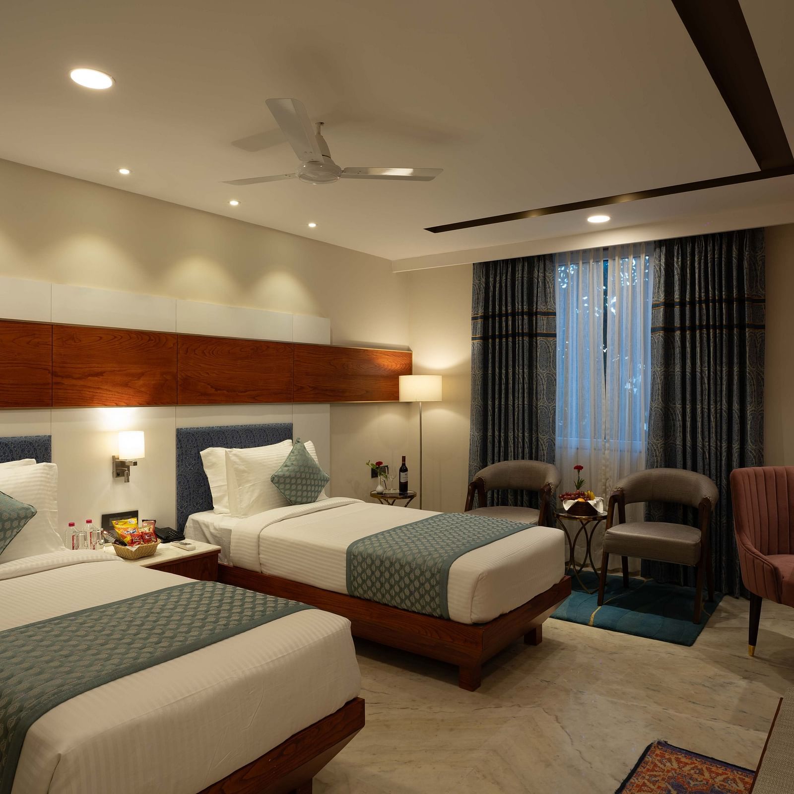 Deluxe Room with twin beds along with a wooden side table with a lamp, wooden table and chair, and two cushioned pieces of furniture at Inde Signature Jaipur