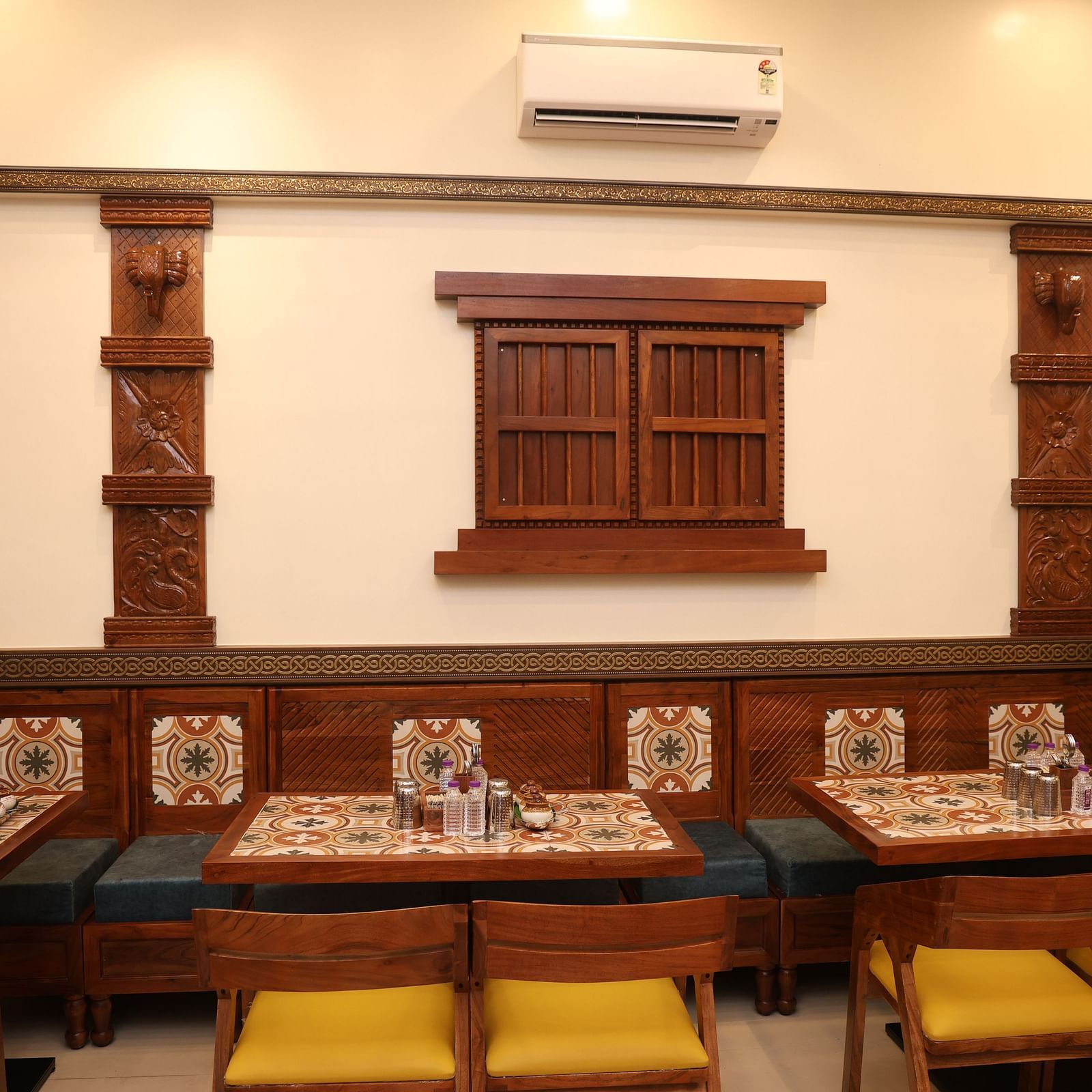 Indoor dining setup with traditional wooden décor, intricately carved pillars, tiled table settings, and cushioned wooden chairs with yellow seats in a warmly lit restaurant.