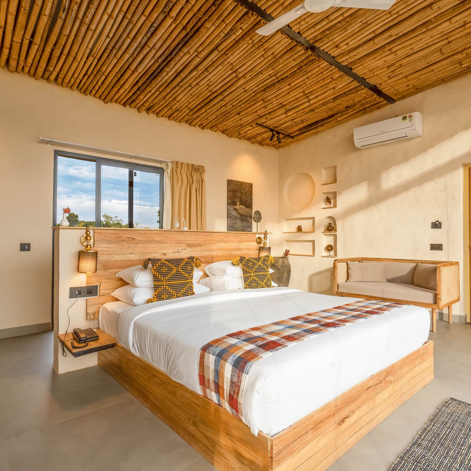 An image of a bedroom featuring a double size, a sofa, ceiling fan, AC, bedside table with lamp, and few decorative elements at Kurja Jawai