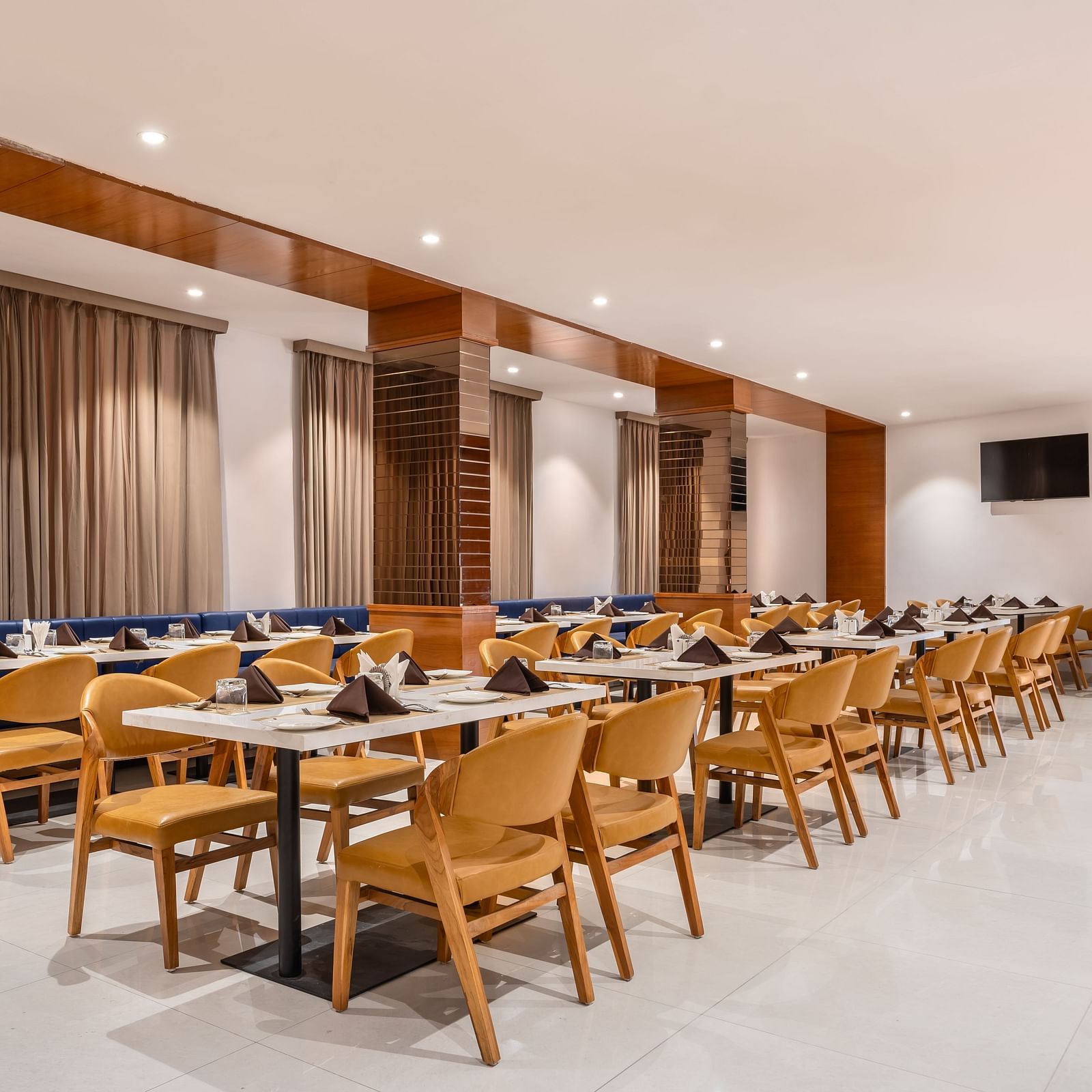 A brightly lit dining area features numerous set tables with chairs and booth seating along the wall - Lords Eco Inn Ambaji.