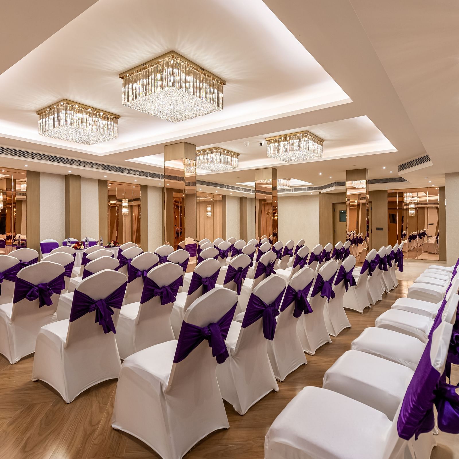 An event space with rows of white chairs decorated with purple fabric, ready for guests. It features a decorative ceiling with hanging lights and mirrored wall accents - Lords Eco Inn Dehradun.