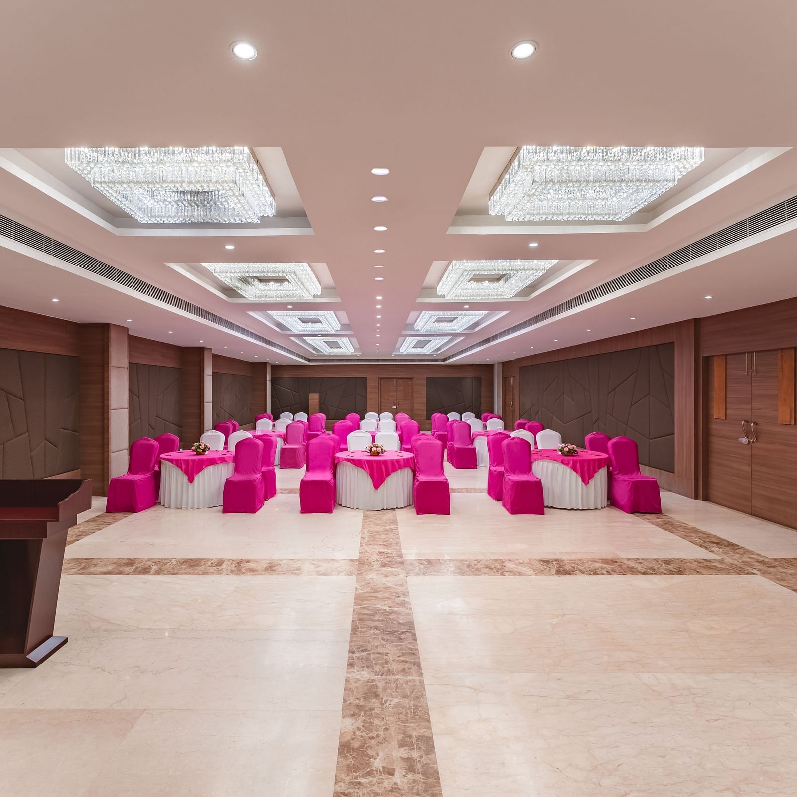 A spacious and well-lit banquet hall at Lords Inn Udaipur is arranged with rows of chairs covered in vibrant pink fabric facing a wooden podium.