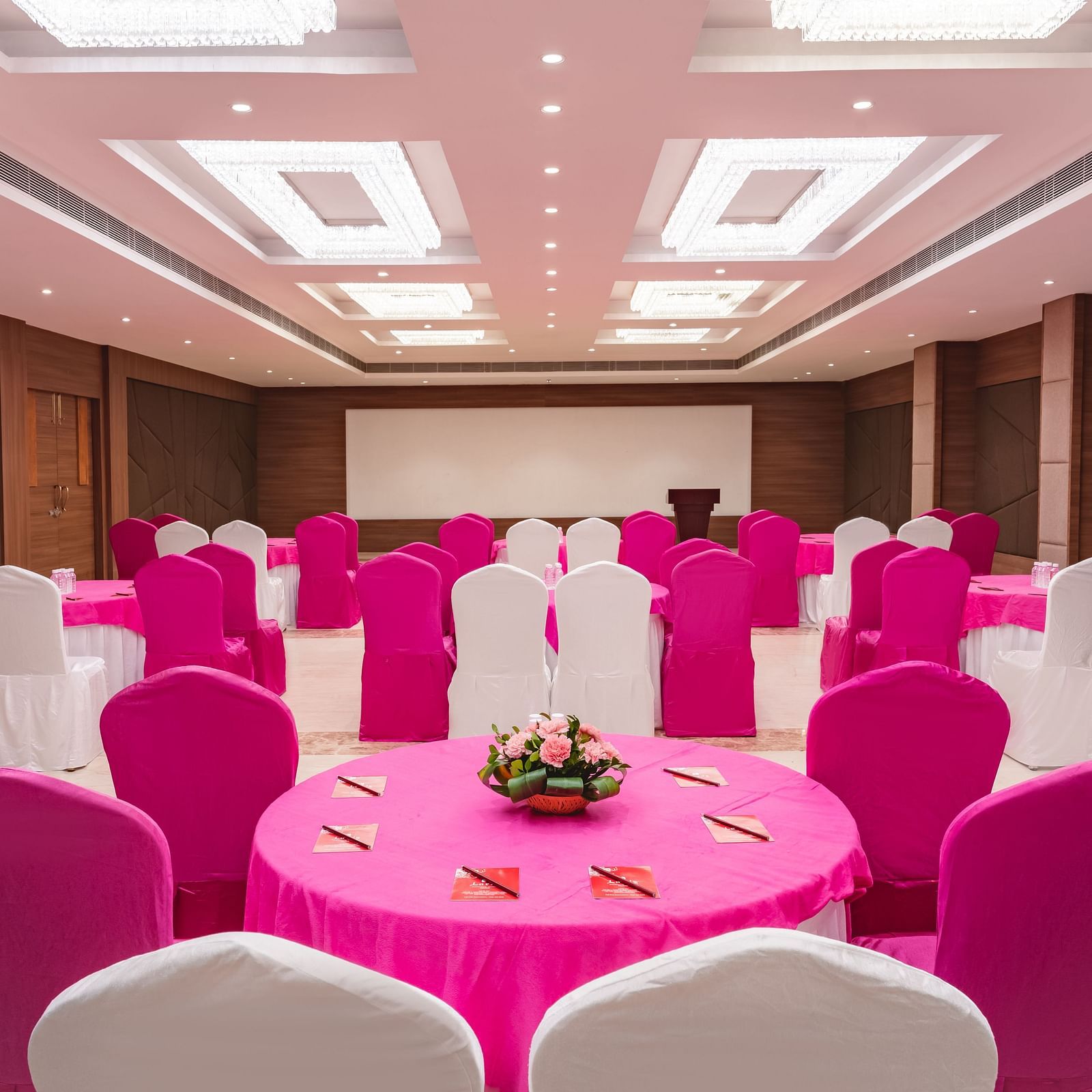 A banquet hall at Lords Inn Udaipur is set for an event with round tables draped in pink, surrounded by chairs in pink and white covers, facing a stage.