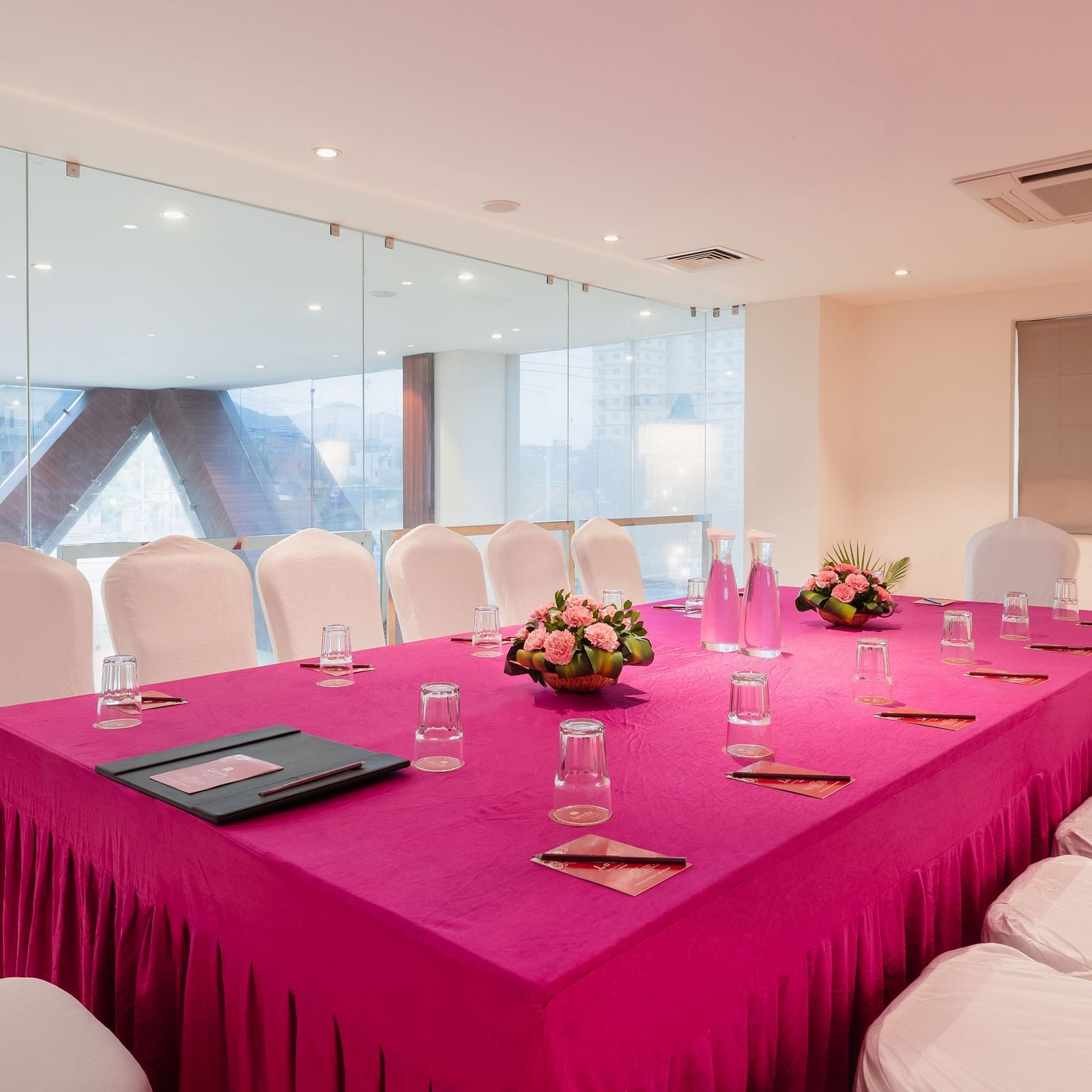 A meeting room at Lords Inn Udaipur is set with a long table covered in a vibrant cloth, surrounded by white chairs and adorned with floral centerpieces and water glasses.