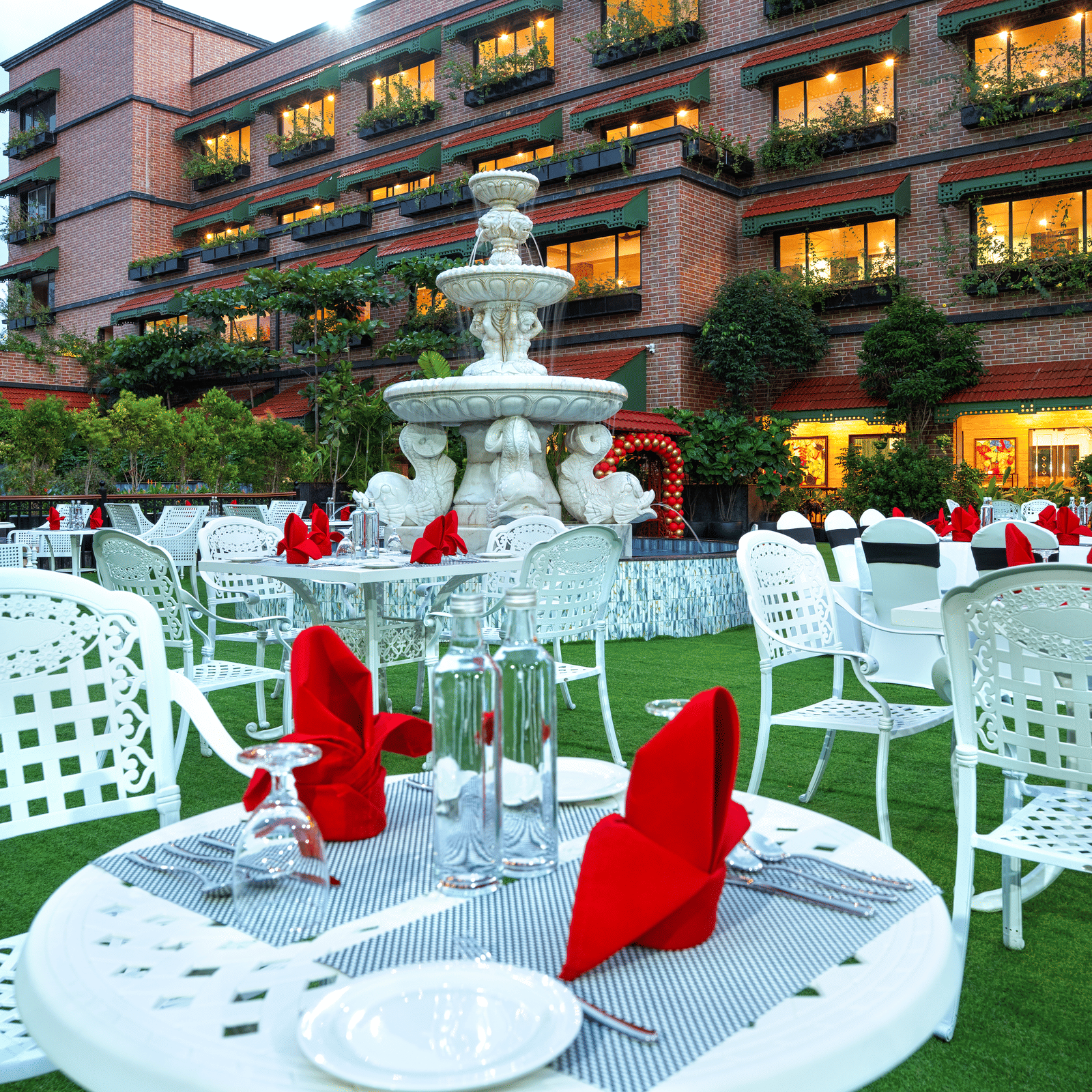 Outdoor event setup on the lawn, showing white lattice tables dressed with red napkins, centred around the fountain at MAYFAIR Bay Resort, Paradeep.