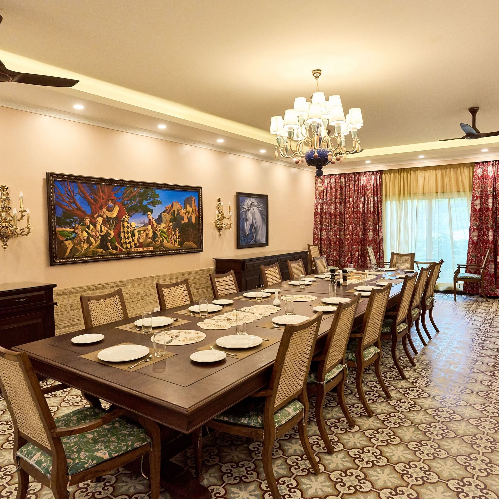 Diagonal view of a large dining table set for a culinary experience, with wall-mounted paintings, a chandelier, and large windows at Mayfair Manor, Alibag