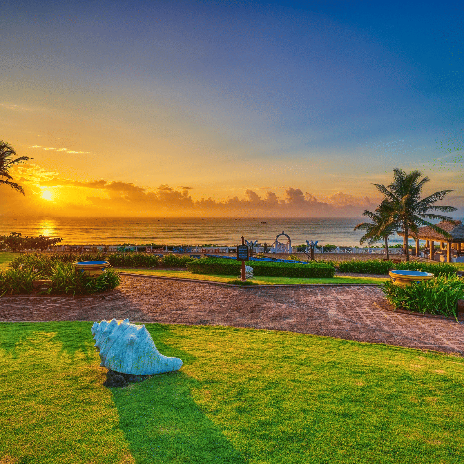 Seafront lawn at sunrise with a large conch-shell sculpture, palm trees, garden pathways and a gazebo at MAYFAIR Palm Beach Resort, Gopalpur-on-Sea.