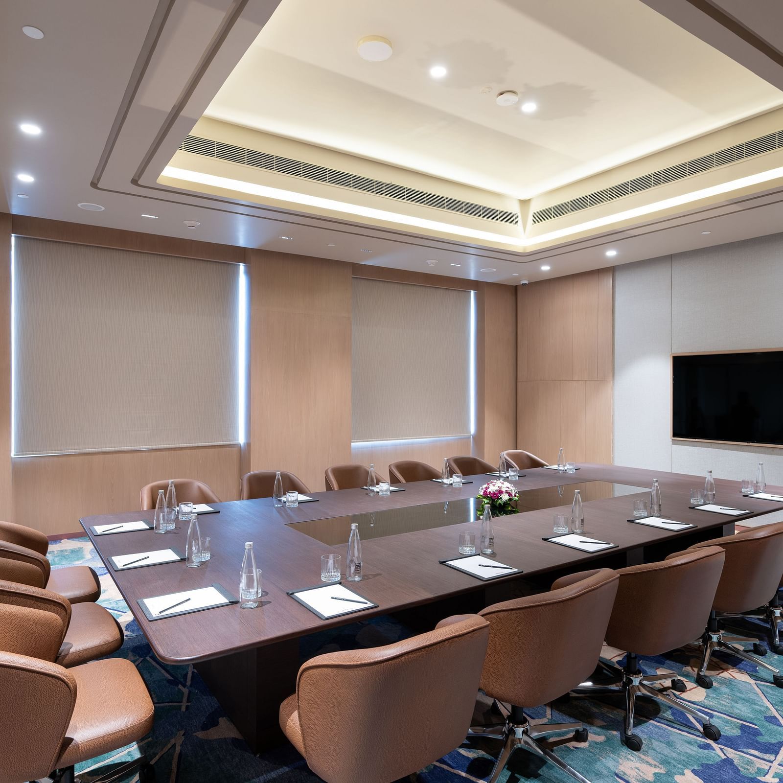Diagonal view of a conference room featuring a projector, large table, and cushioned chairs at Merlis Hotel, Coimbatore.