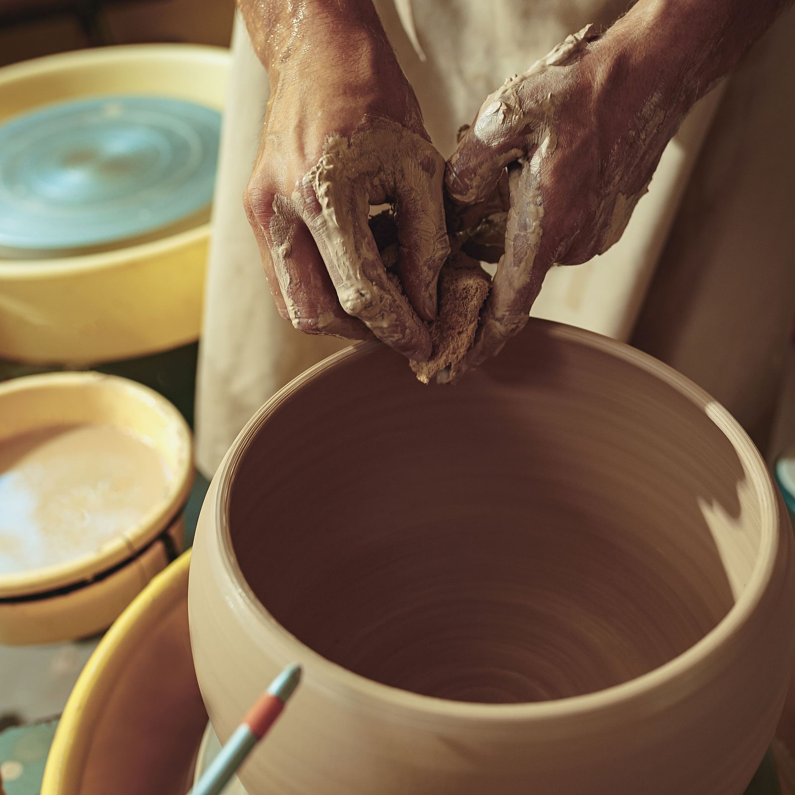 Potter shaping a clay vase on a spinning wheel, with hands covered in clay and tools visible on the worktable.