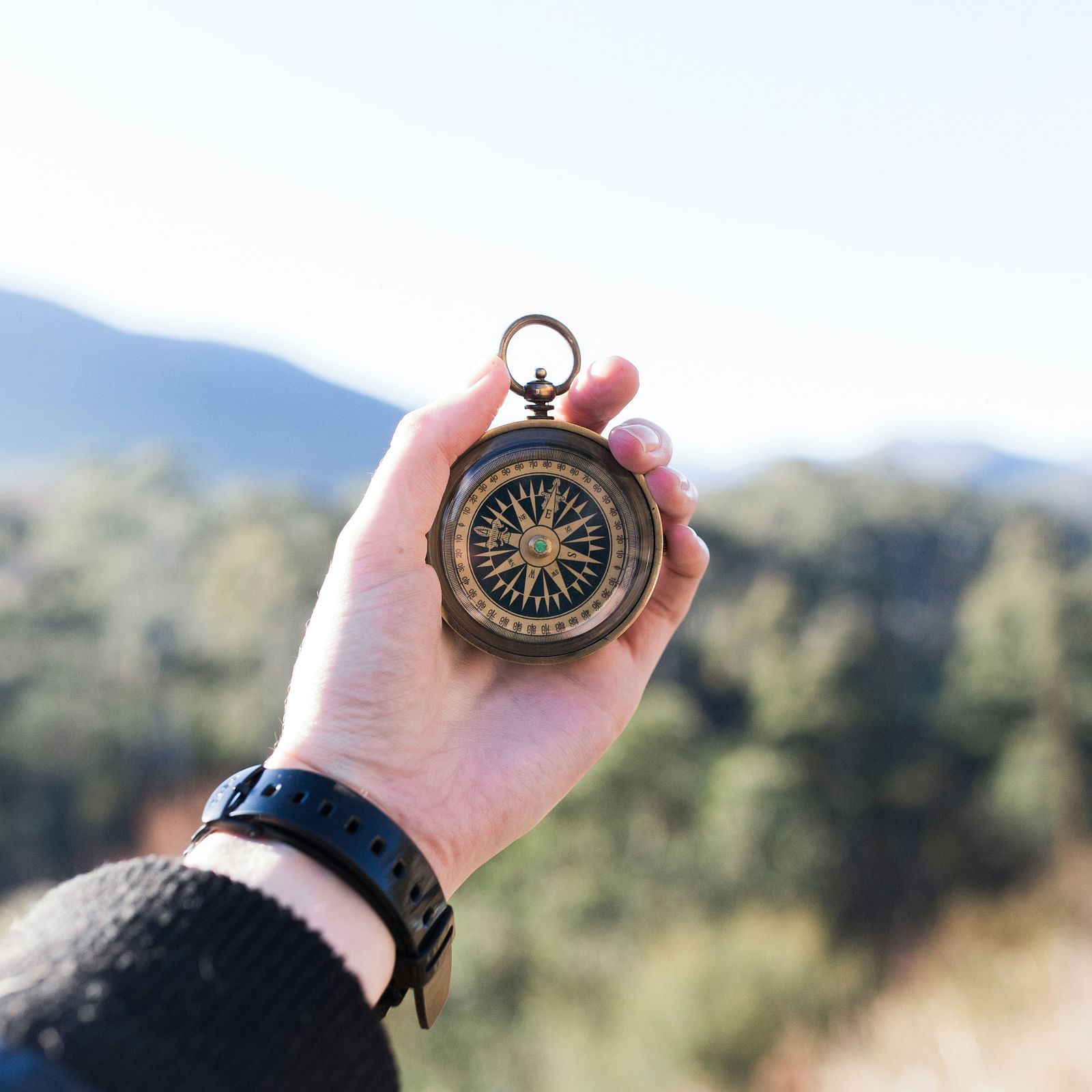 The image shows a person holding a vintage brass compass with a scenic mountainous background.