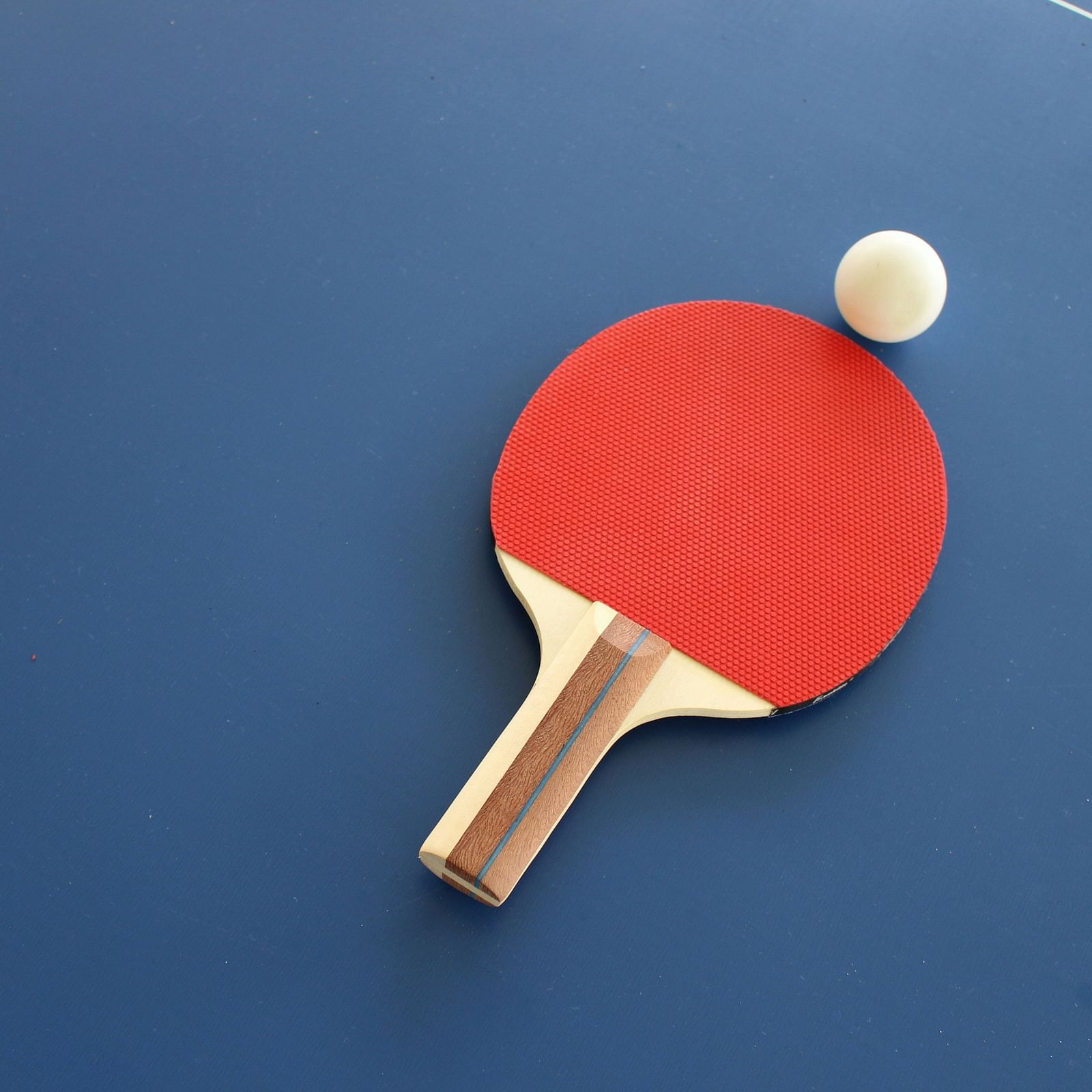 A close-up view of a red table tennis paddle and a white ball on a blue table, ready for a game of ping pong.