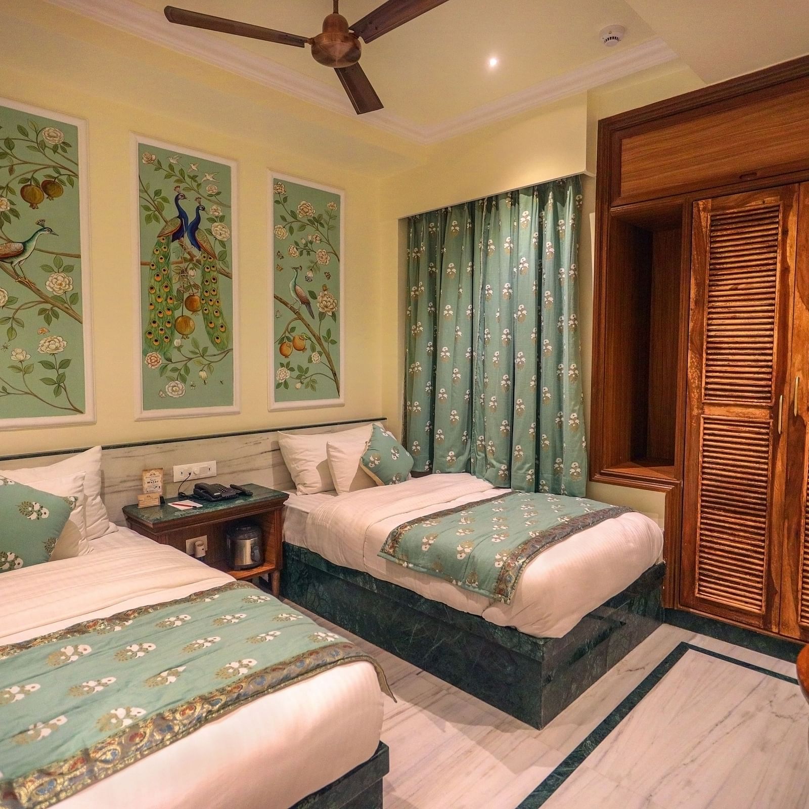 Diagonal view of the spacious room with twin beds placed side by side, a wooden bedside table in the middle, three photo frames above the headboard, and wooden storage | Panna Vilas Udaipur