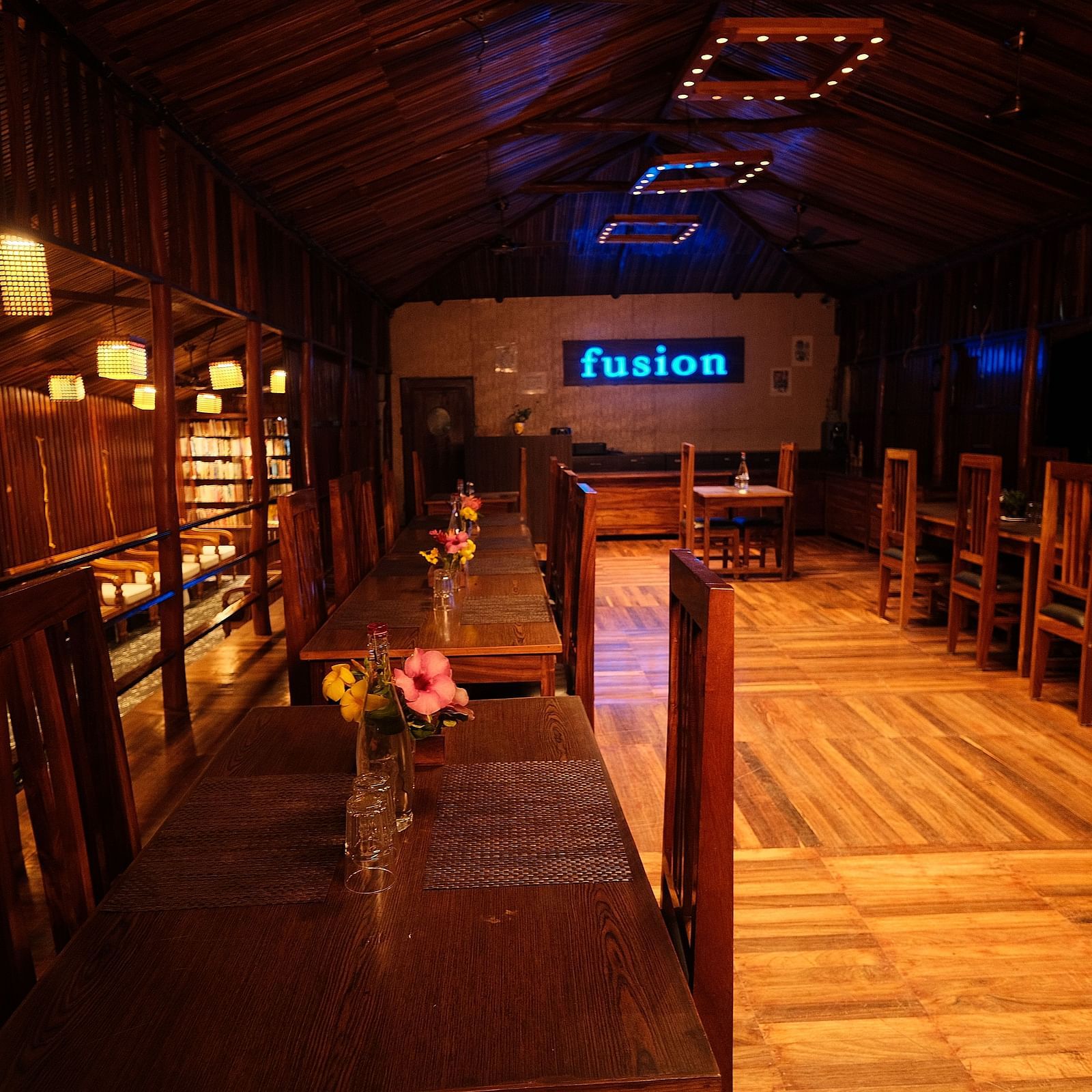 A wooden interior is illuminated by hanging lamps, highlighting the seating and shelving arrangements | Fusion Cafe and Multi-cuisine | RainForest Resort, Port Blair, Andaman and Nicobar Island