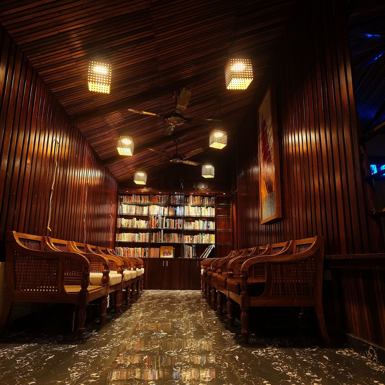 A warmly lit wooden interior shows seating areas and shelves, with hanging lamps casting a soft glow | Fusion Cafe and Multi-cuisine | RainForest Resort, Port Blair, Andaman and Nicobar Island