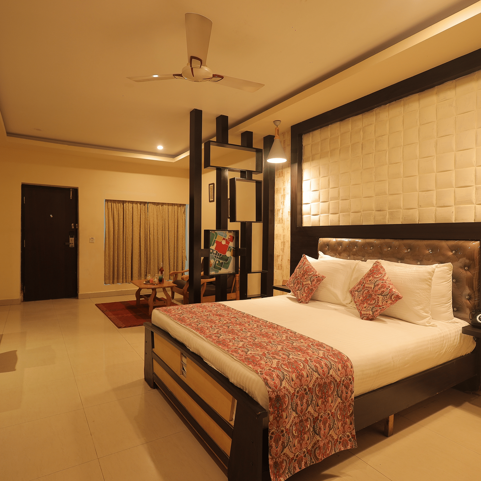 An image of a bedroom with bedside table, a set of table and chairs, flat TV screen, and AC- Sathyam Grand Resort