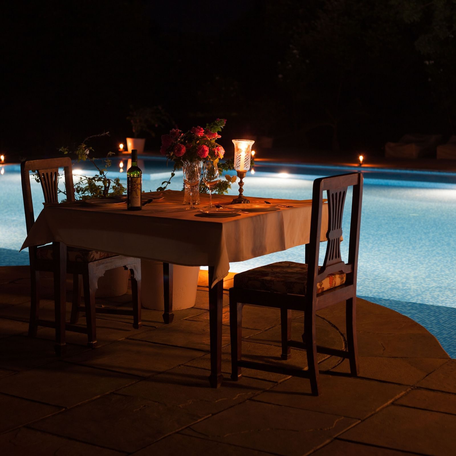 A candlelit dining table set for two by a pool at night, with a dark background- Shahpura Bagh.