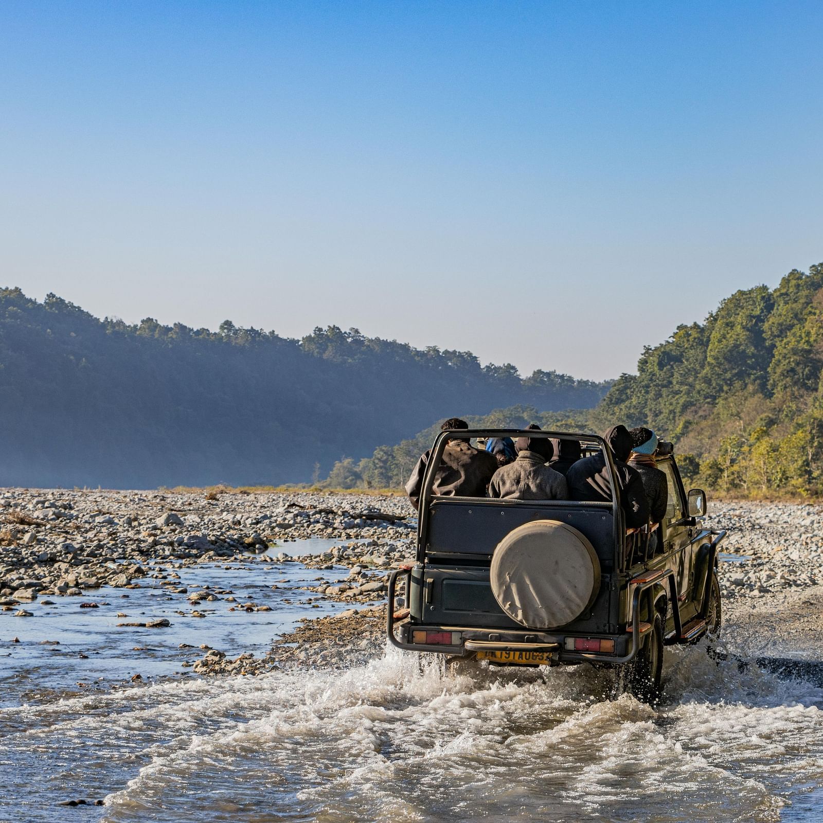 A jeep full of people drives through a shallow, rocky riverbed in Jim Corbett National Park, surrounded by forested hills.