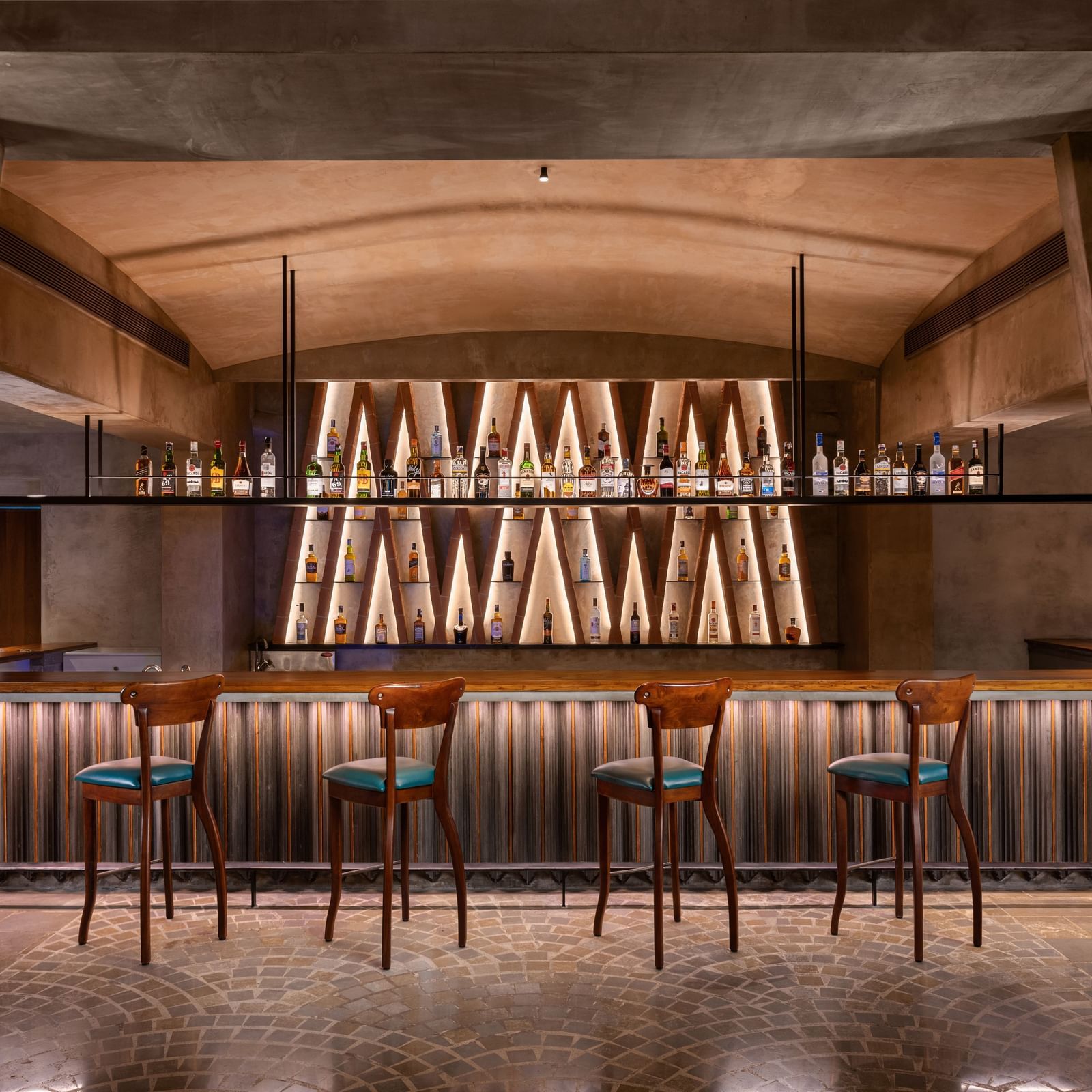 A dimly lit bar with a long counter, several bar stools, and shelves of bottles illuminated from behind, at Shilton International, Kochi.