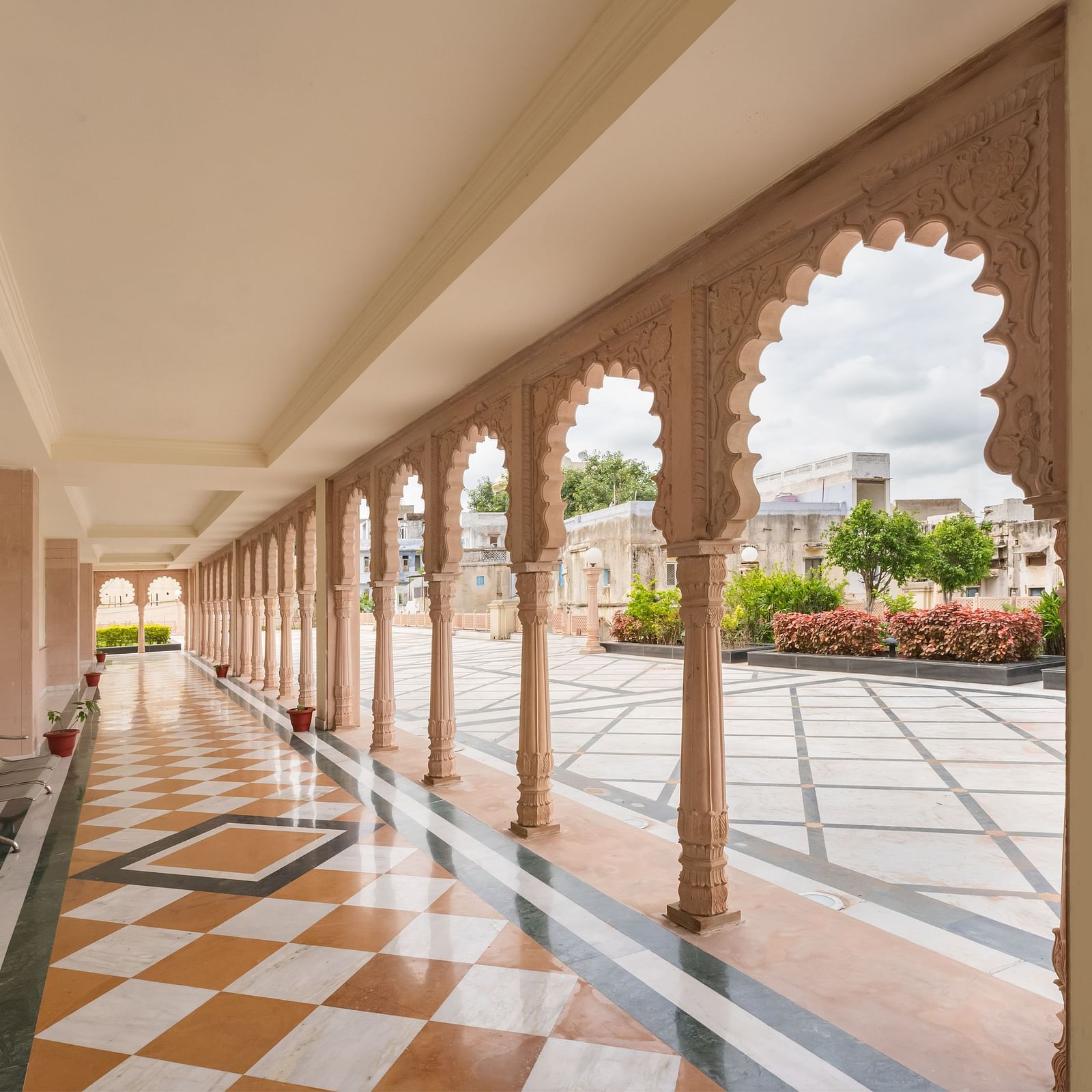 A covered outdoor corridor at Shri Vallabh Vilas Lords Plaza Nathdwara, with arched stone columns and checkered flooring, opening to a garden area.