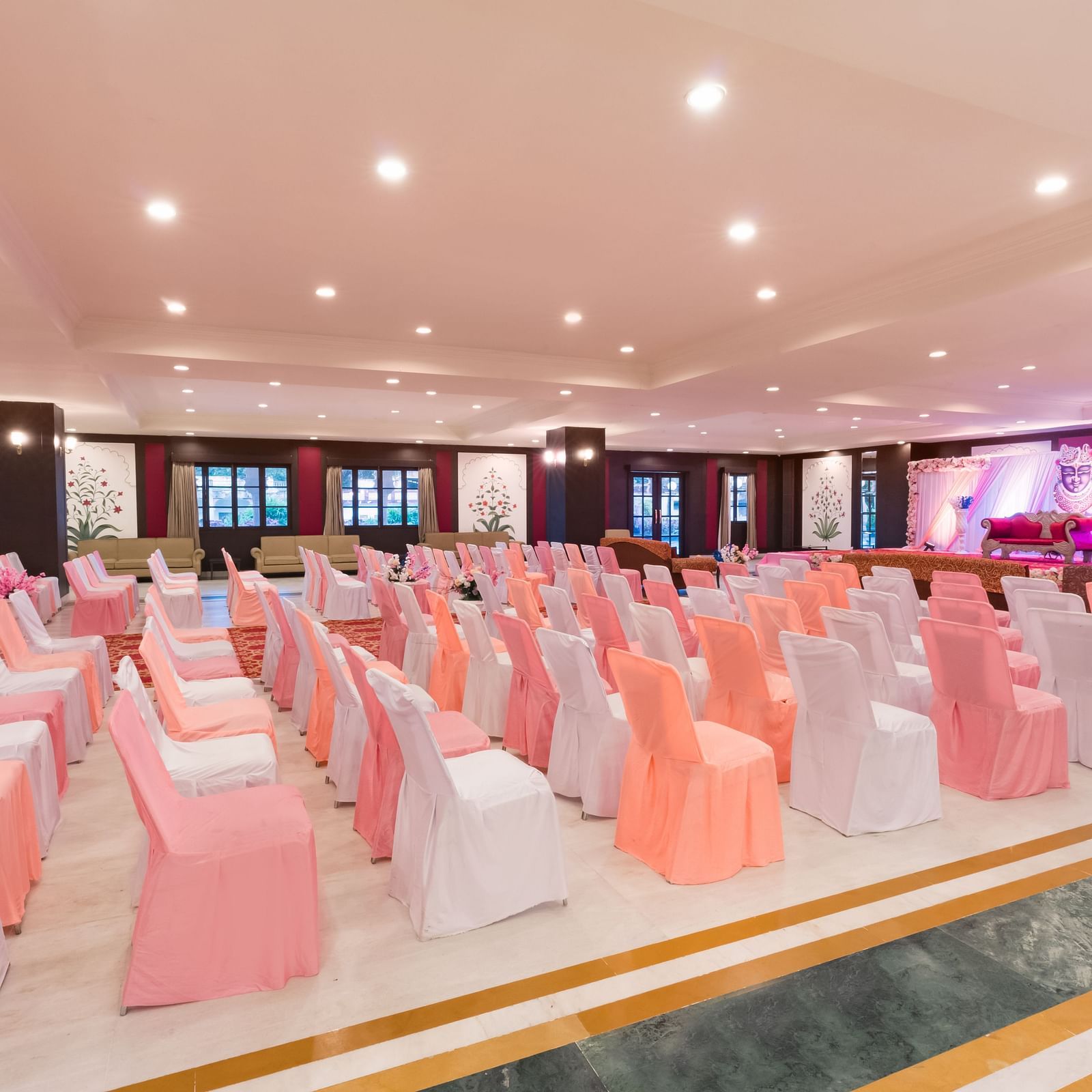 Rows of banquet chairs in white and peach covers arranged for an event in a spacious well-lit Raj Mahal at Shri Vallabh Villas Lords Plaza Nathdwara.