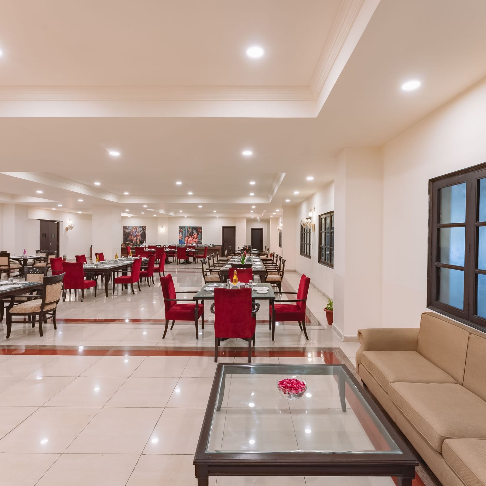 An indoor well lit spacious dining area at Shri Vallabh Vilas Lords Plaza Nathdwara, featuring red chairs, tiled floor and a seating lobby area with windows.