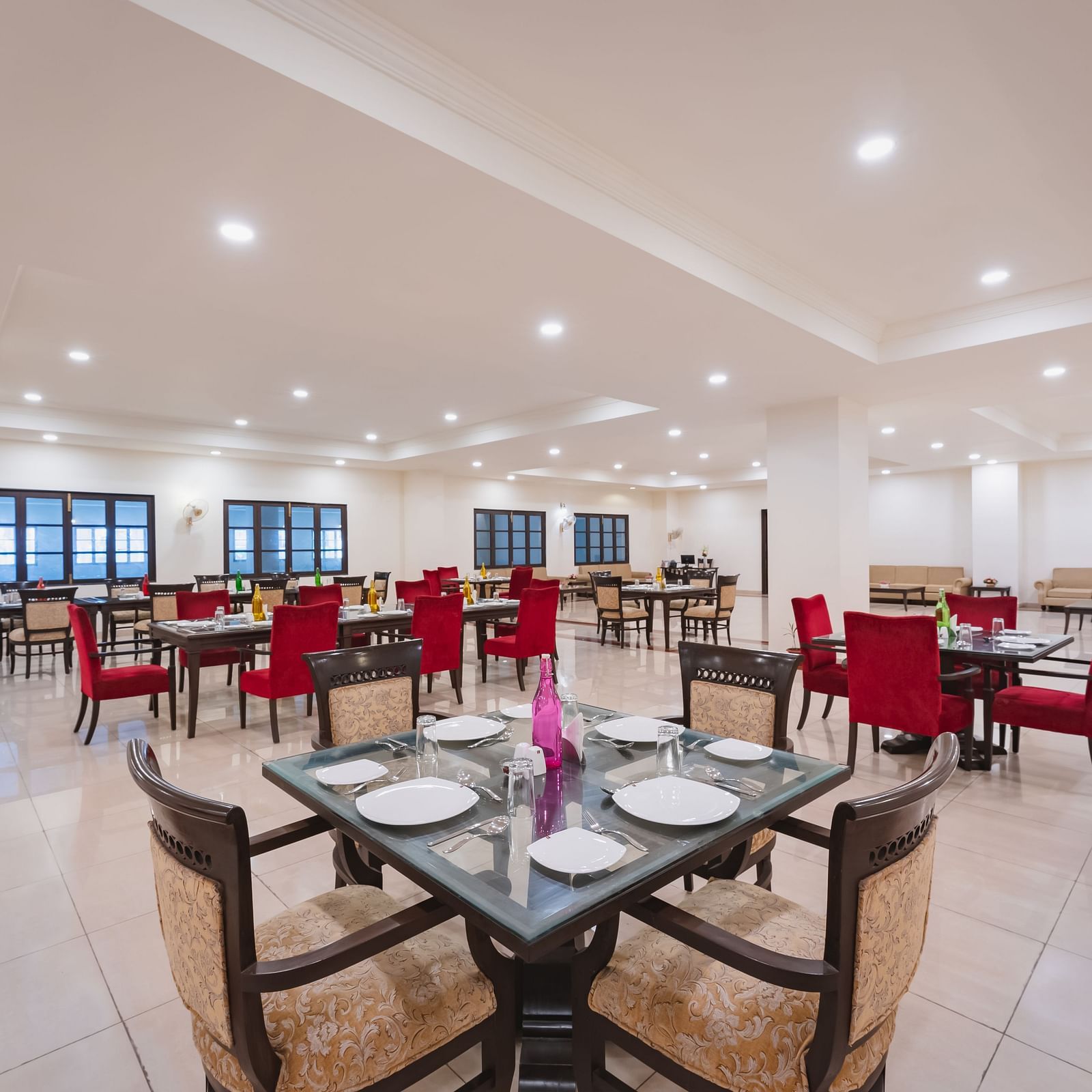 An indoor dining area Shri Vallabh Vilas Lords Plaza Nathdwara featuring red and beige coloured chairs with wooden furnishings, clean tile floor and rows of windows.